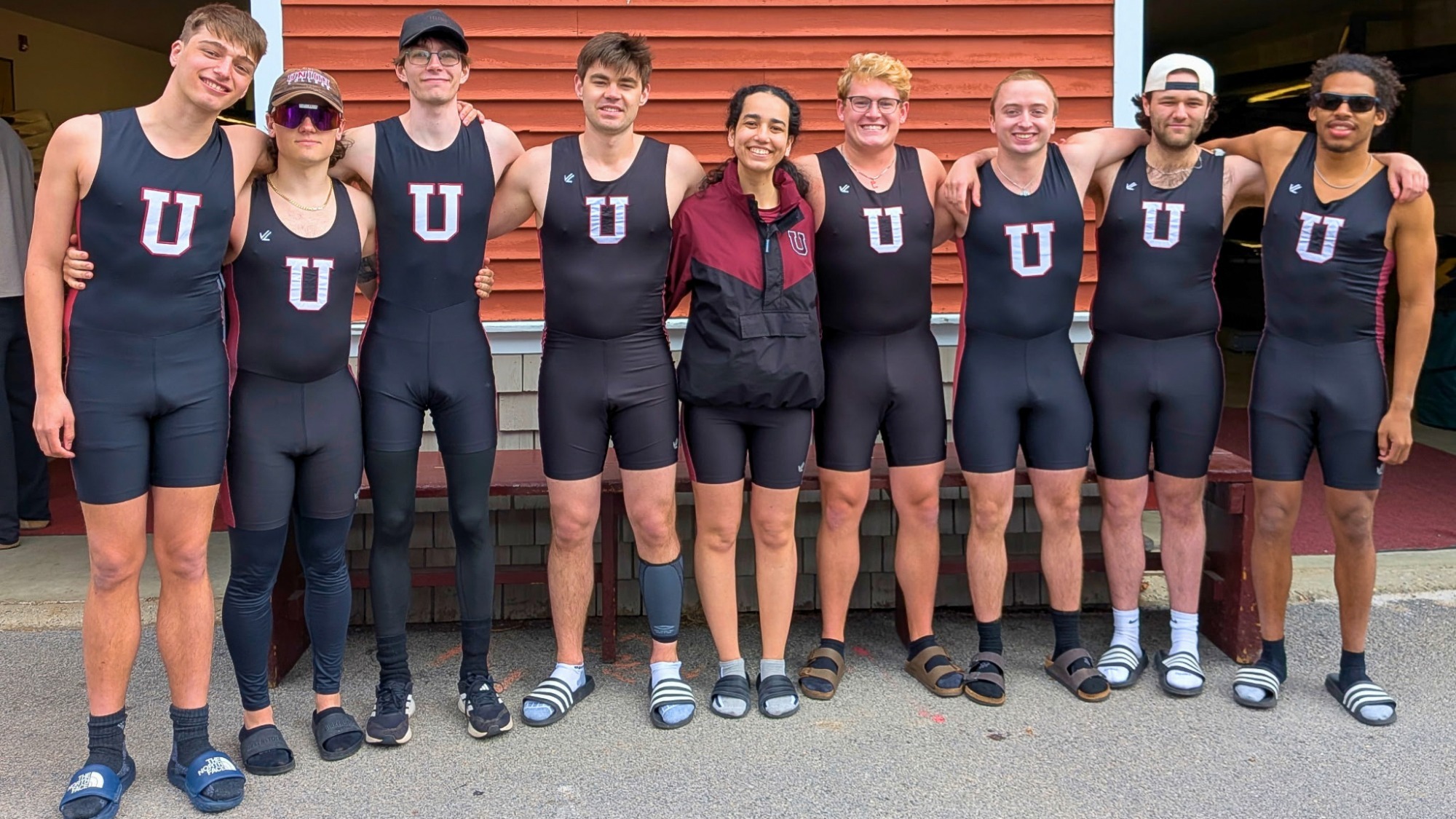 Nine crew athletes standing arm-in-arm in front of a reddish-orange building, smiling at the camera and wearing all-black with a white Block-U on the front of their gear
