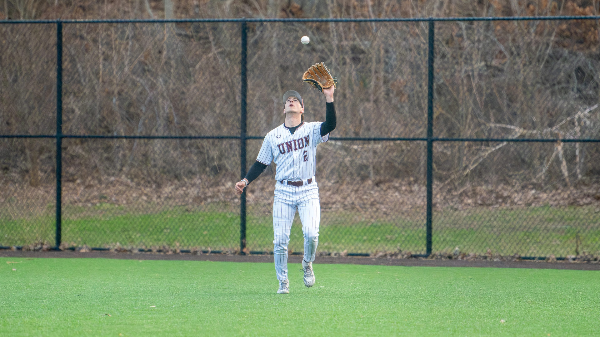 a baseball player wearing a white jersey with garnet pinstripes, holding his left arm up to catch a baseball that is about to fall into his glove