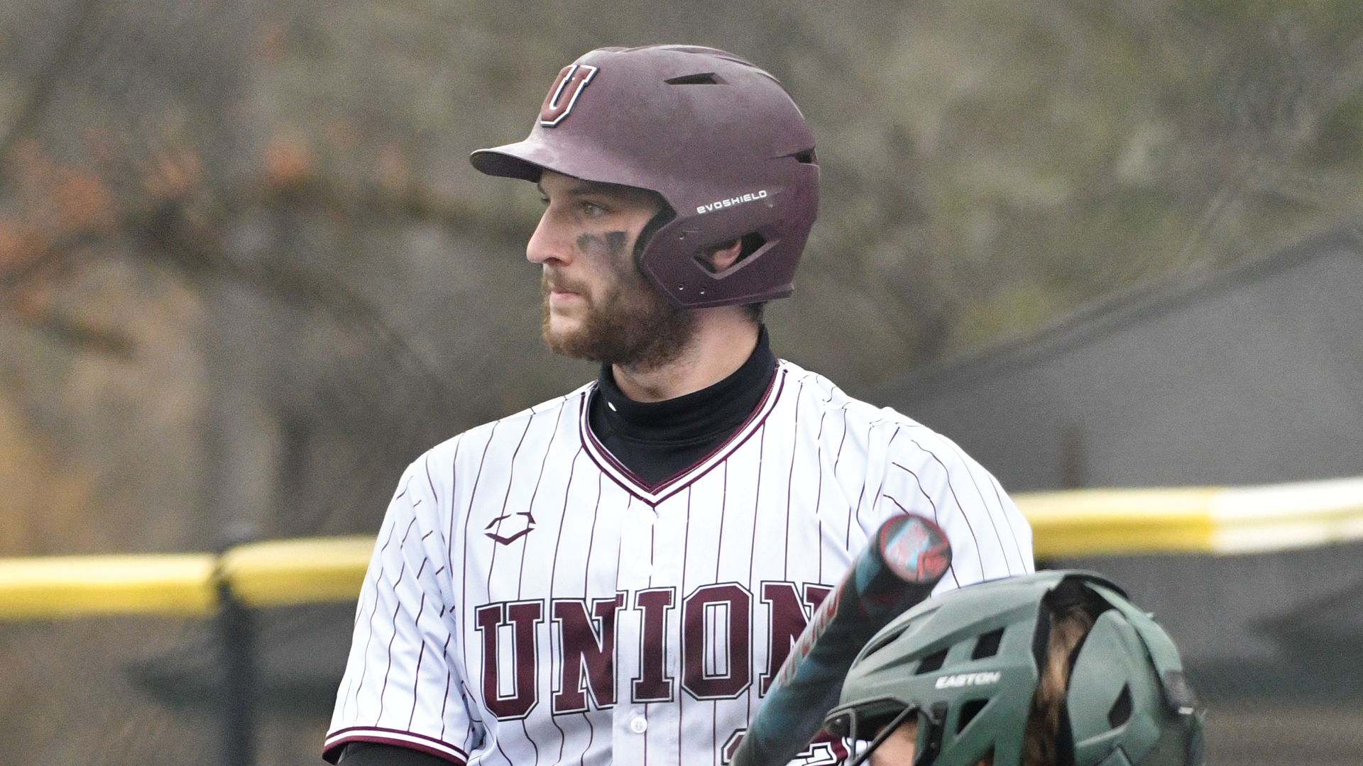 a baseball player looking out on the field wearing a garnet-colored batting helmet and a white jersey with thin, garnet pinstripes