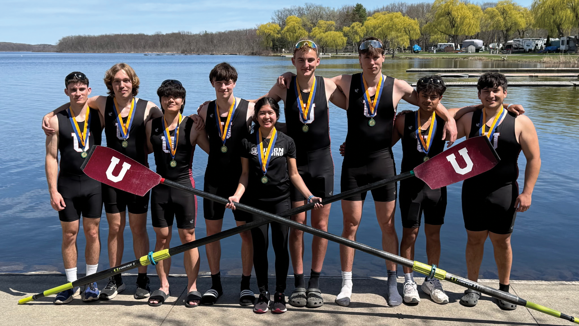 a men's crew team standing arm-in-arm in front of calm waters