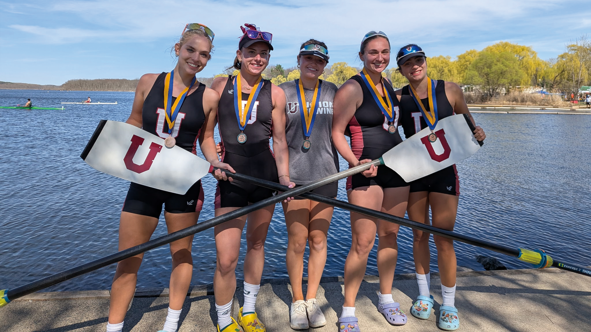 five women's crew student-athletes holding intersecting oars, posing in front of water
