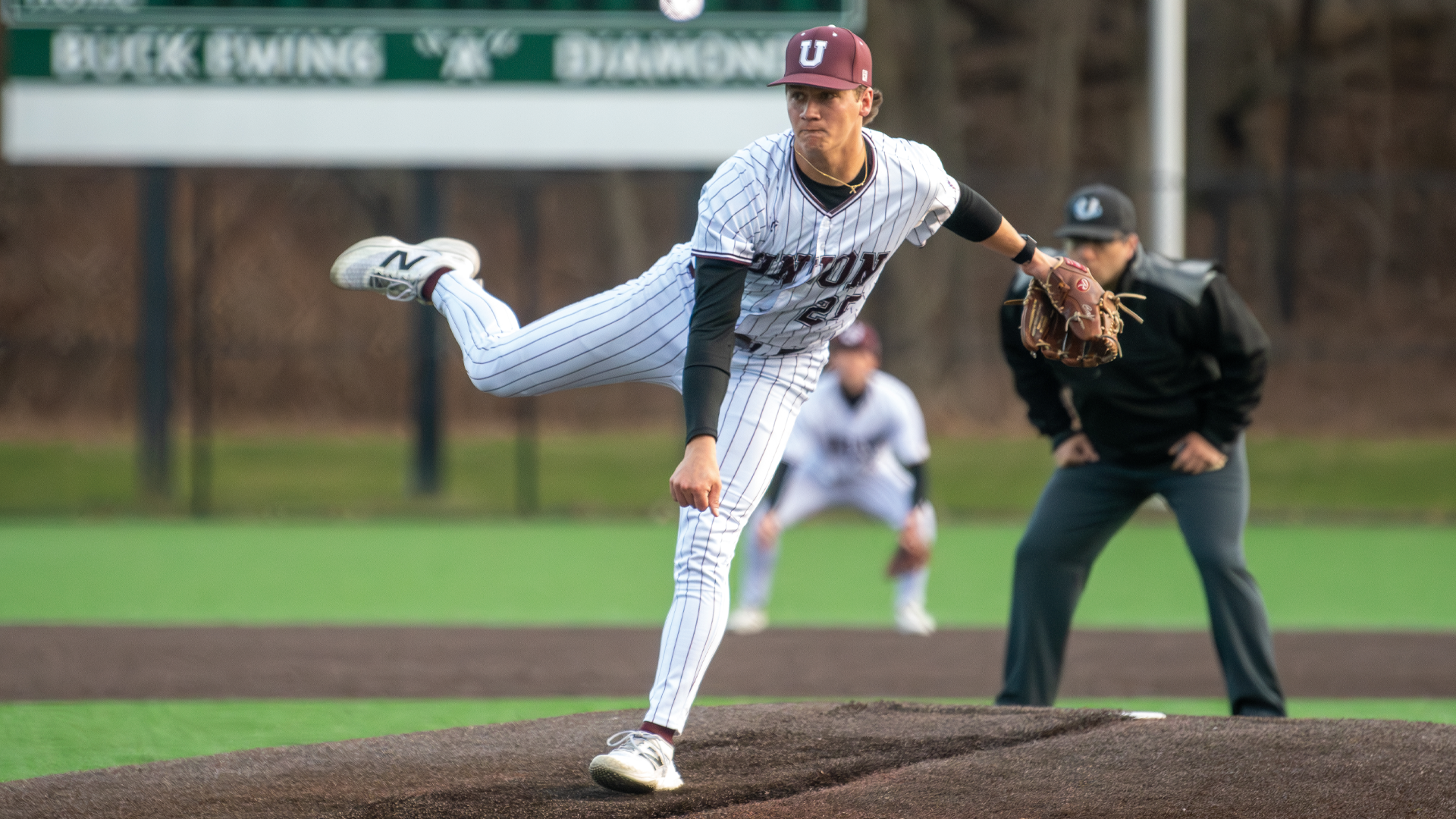 baseball pitcher planted on his left leg during the follow through of his pitch, wearing a white jersey and garnet-colored hat as the sun sets, creating a golden glow on the player's profile