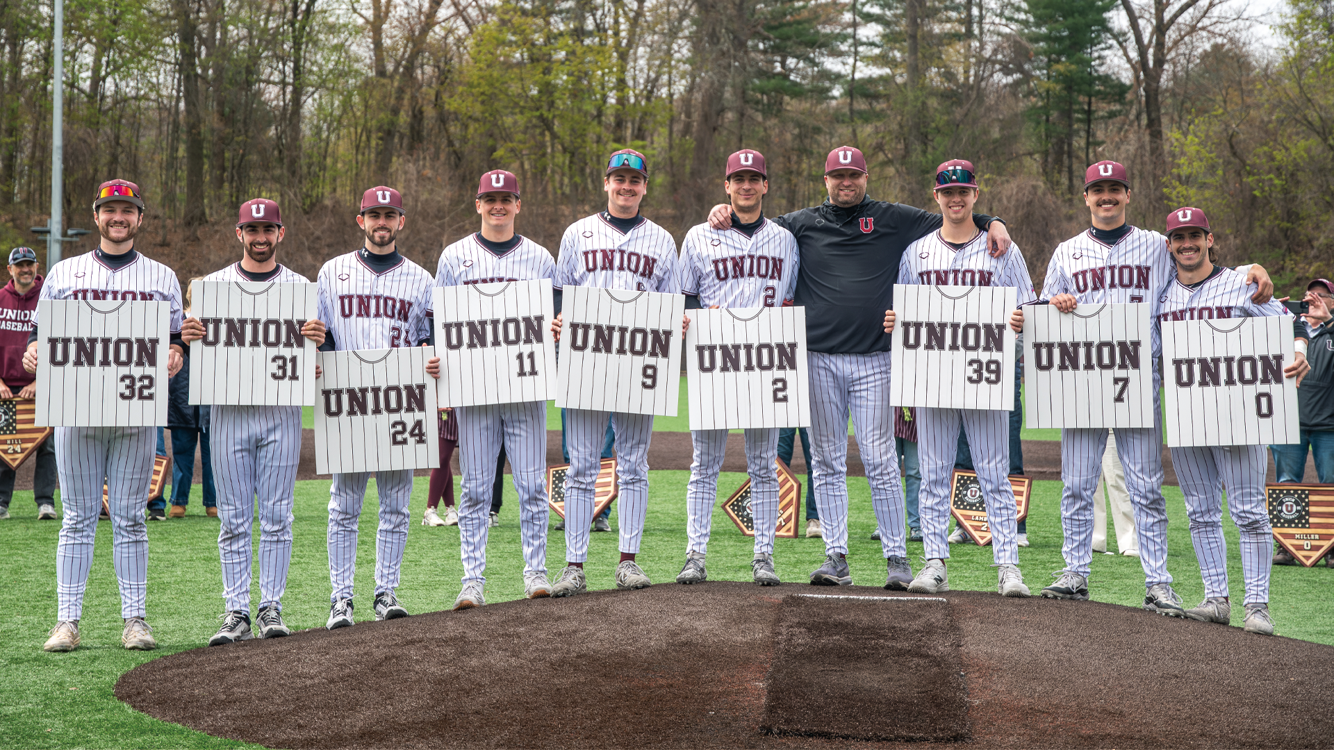 nine baseball players and their coach standing along the pitcher mound, smiling at the camera and holding square paintings of their jerseys