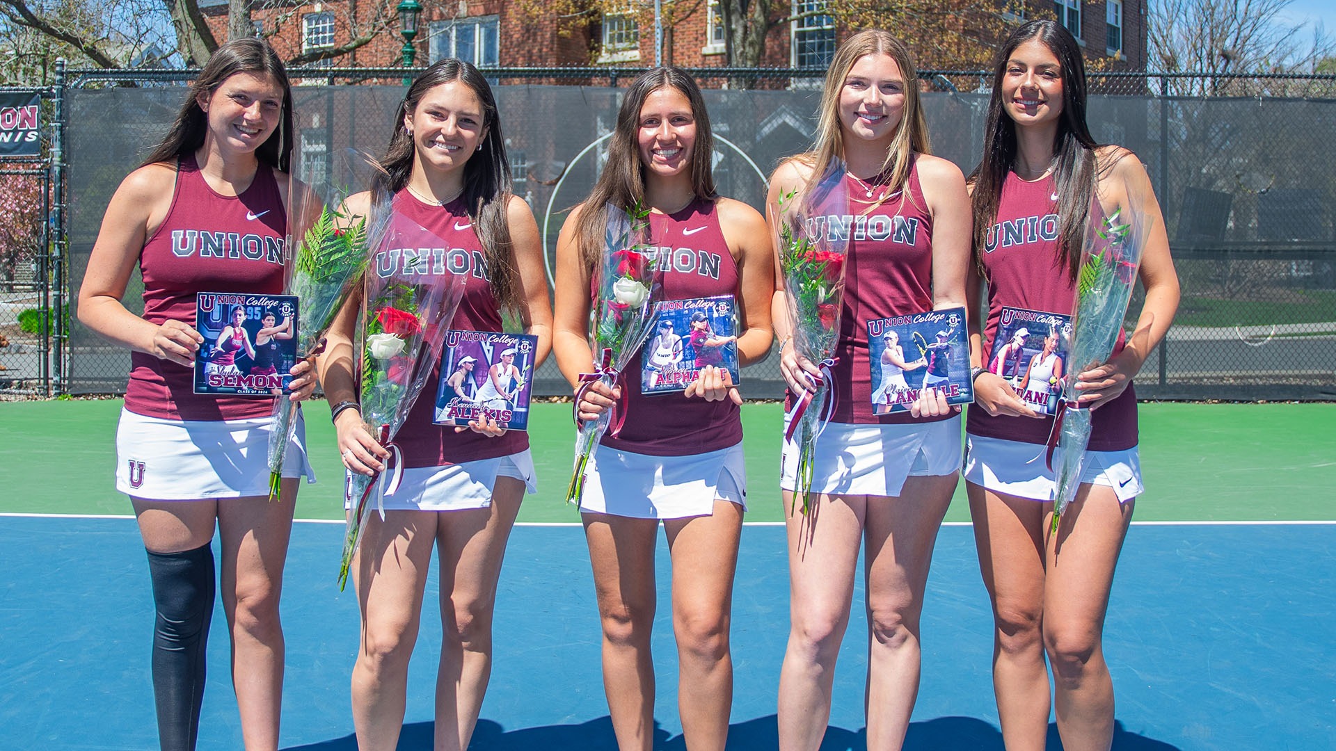 five women's tennis players standing side-by-side on a blue tennis court, holding flowers and a collage of photos of themselves playing tennis