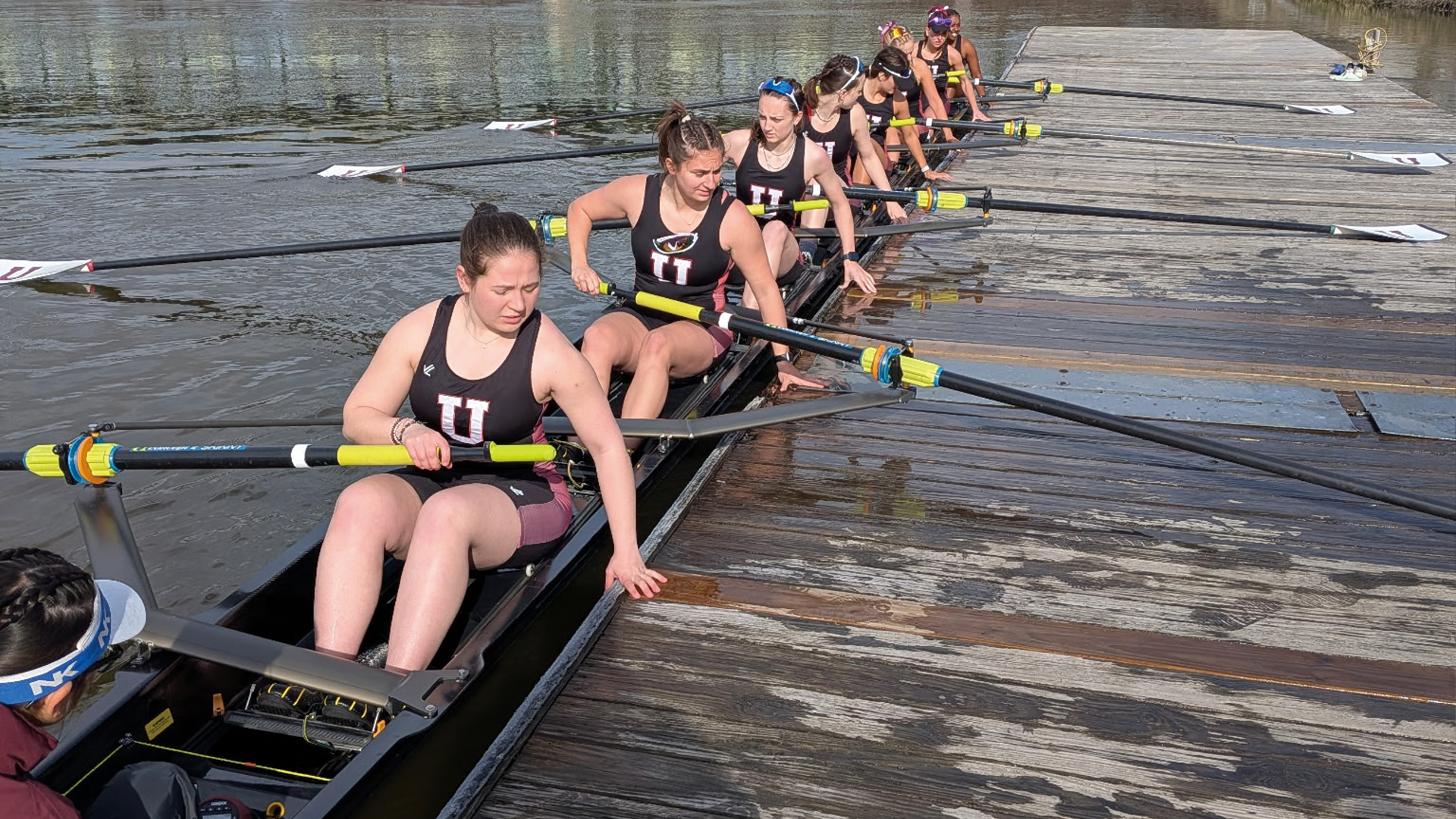 a women's crew team in a boat, all pushing off a wooden deck to their left