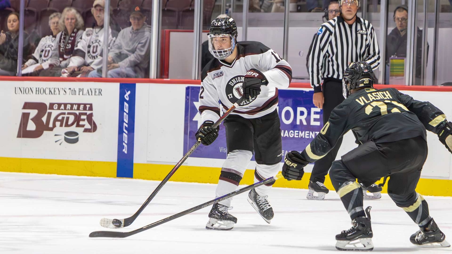 a men's hockey player making a pass with a defender wearing all black skating backwards in front of him