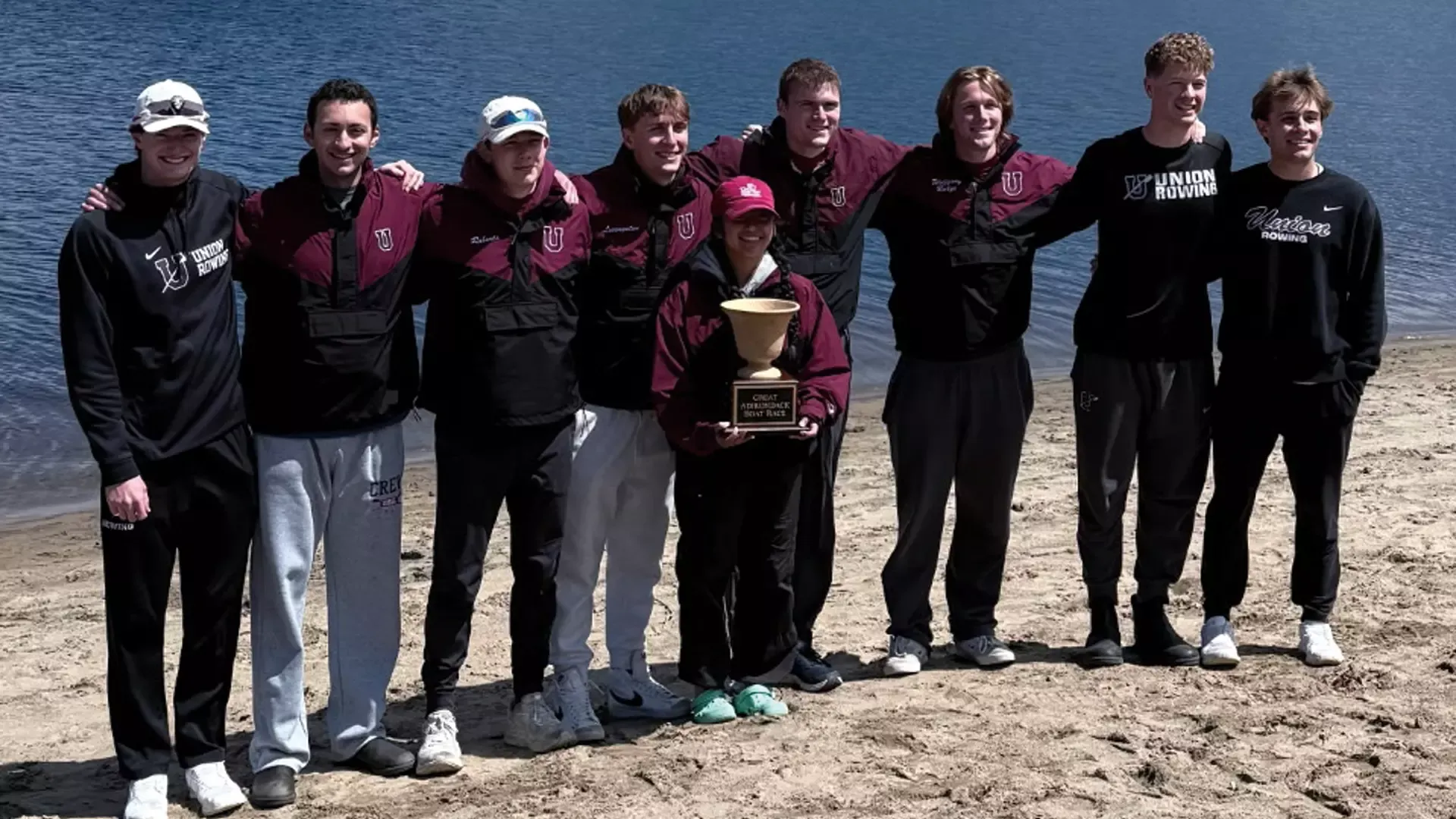 a men's crew team holding a trophy on the shore of a lake