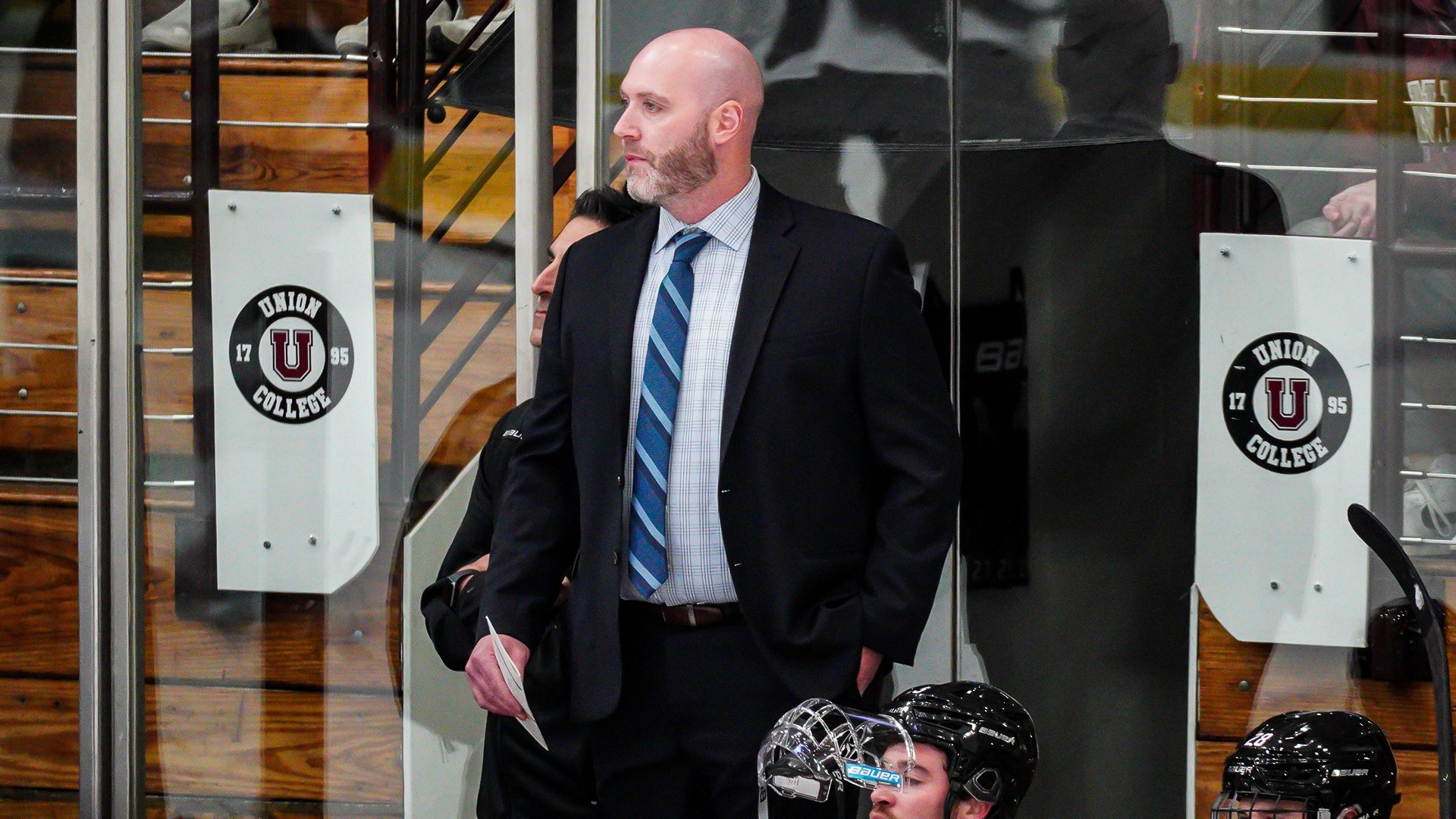 A hockey coach stands on the bench looking at the ice with players seated in front of him.