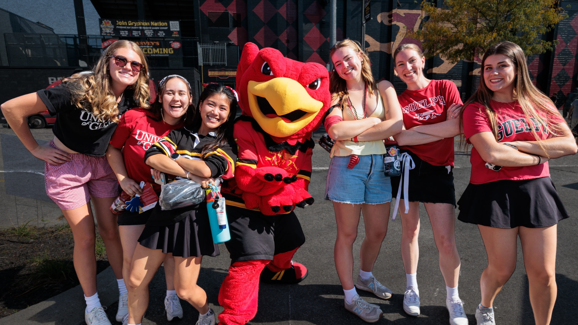 Six people smiling and posing with the Gryphon mascot in front of Alumni Stadium at Homecoming wearing fan-gear. 