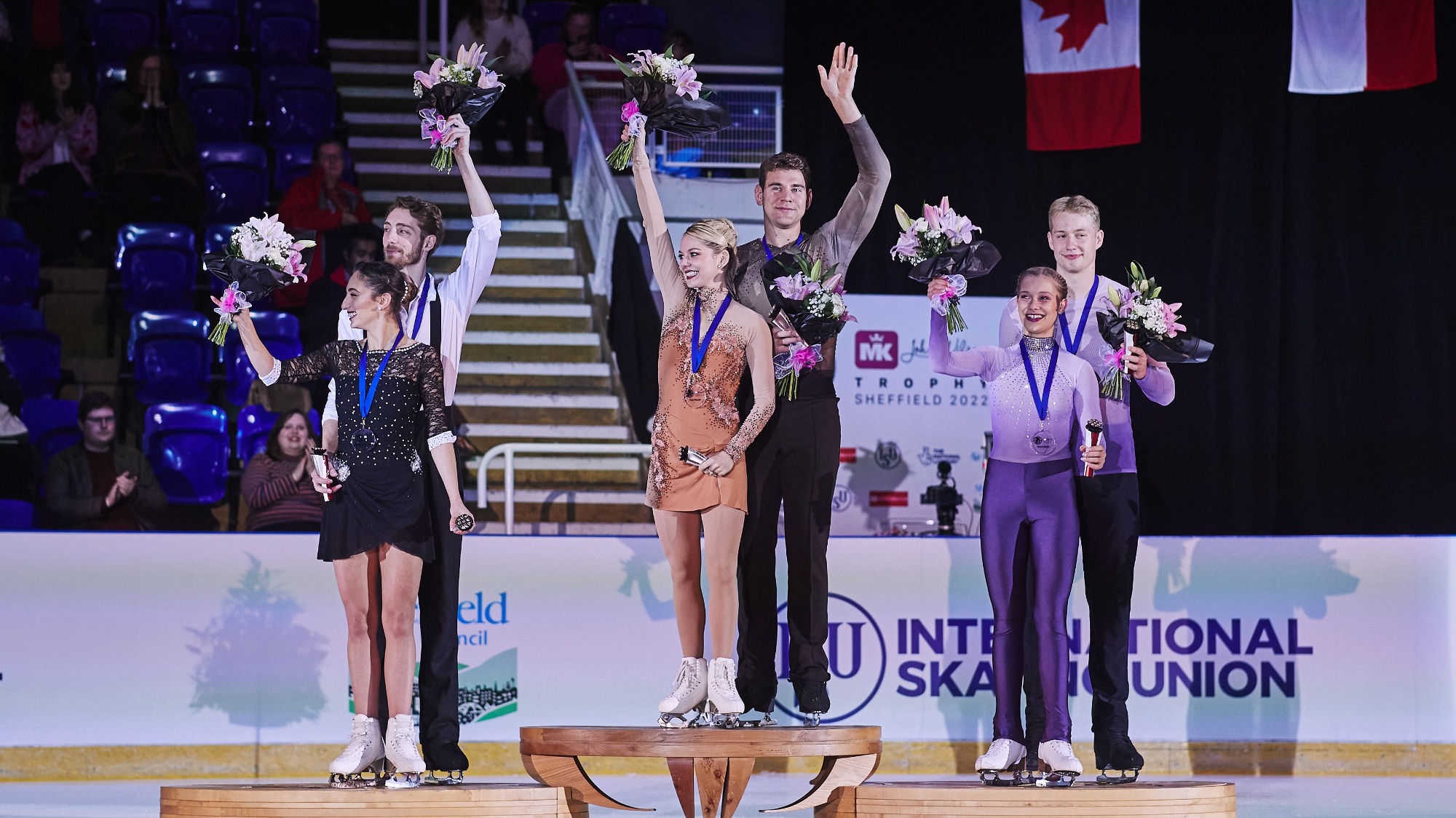 Alexa Knierm and Brandon Frazier stand atop the podium and wave, gold medals around their necks at the MK John Wilson Trophy. Next to them on the podium are Sara Conti and Niccolo Macii of Italy and Letizia Roscher and Luis Schuster of Germany.