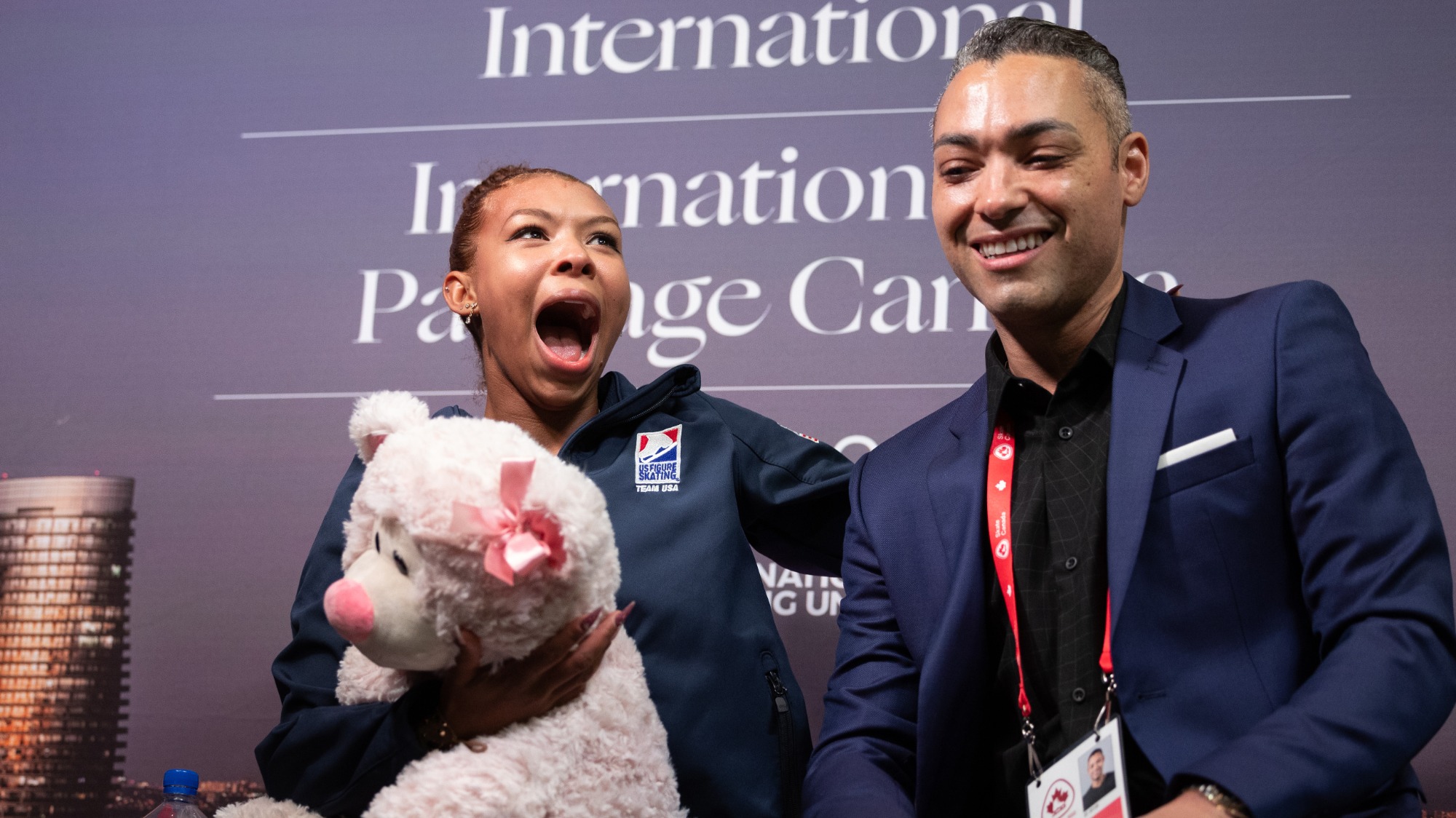 Starr Andrews looks in shock while holding a teddy bear and sitting next to her coach Derrick Delmore in the kiss and cry after hearing her scores at Skate Canada International.