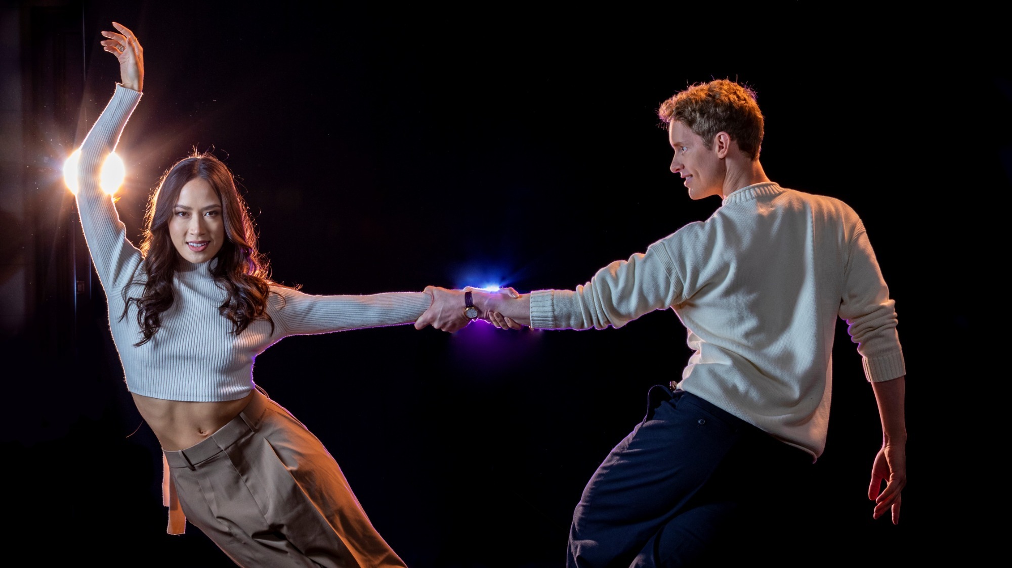 Madison Chock and Evan Bates during a photoshoot again a dark backdrop, Evan holding up Madison as they grip each other's arm and Madison looking straight into the camera while hitting an elegant pose.