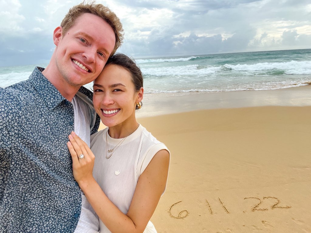 Evan Bates and Madison Chock, with an engagement ring, pose in front of the ocean with the date6.11.22 written in the sand.