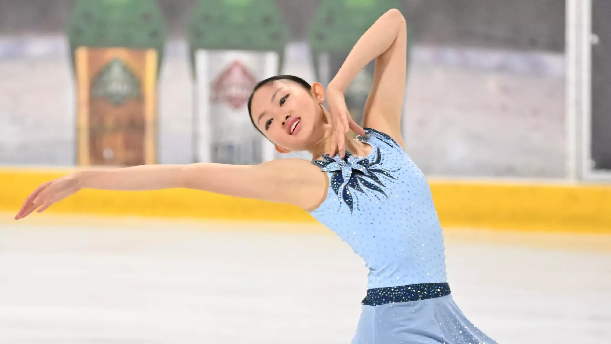 Audrey Lu, in a light blue and navy dress with a flower at the top, looks over her right shoulder with her right arm extended in a pose.