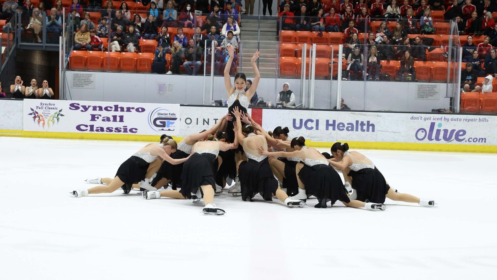 Photo of the Skyliner junior team competing. They are all wearing skating costumes with sparkly silver tops and black skirts. One athlete stands in the middle of the group with her hands held above her head. All of her teammates surround her on their knees as if they are all bowing to her. 