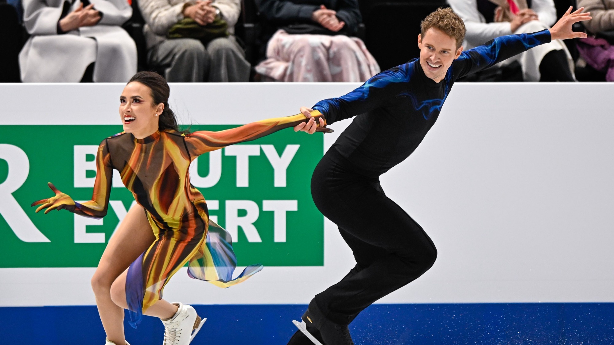 Madison Chock and Evan Bates perform their free dance. She wears a dress that looks like flames, skating to the left and he holds her hand and skates behind her, facing to the right. Both are smiling. He wears all black with blue flames on the arms.