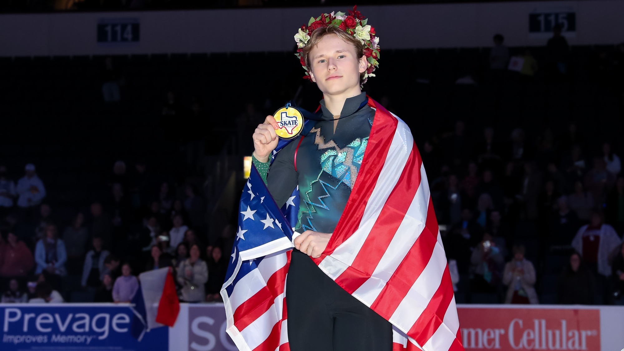 Ilia Malinin, draped in the U.S. flag, holds up his Skate America gold medal while standing on the ice. He wears a flower crown.