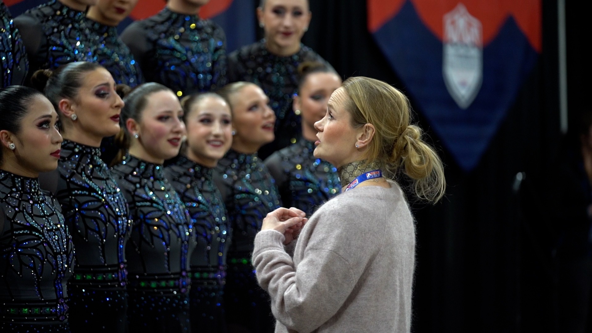 Saga Kranz, wearing a grey cardigan, faces and speaks to her team, the Haydenettes, who stand in the Kiss and Cry at the 2023 U.S. Synchronized Skating Championships.