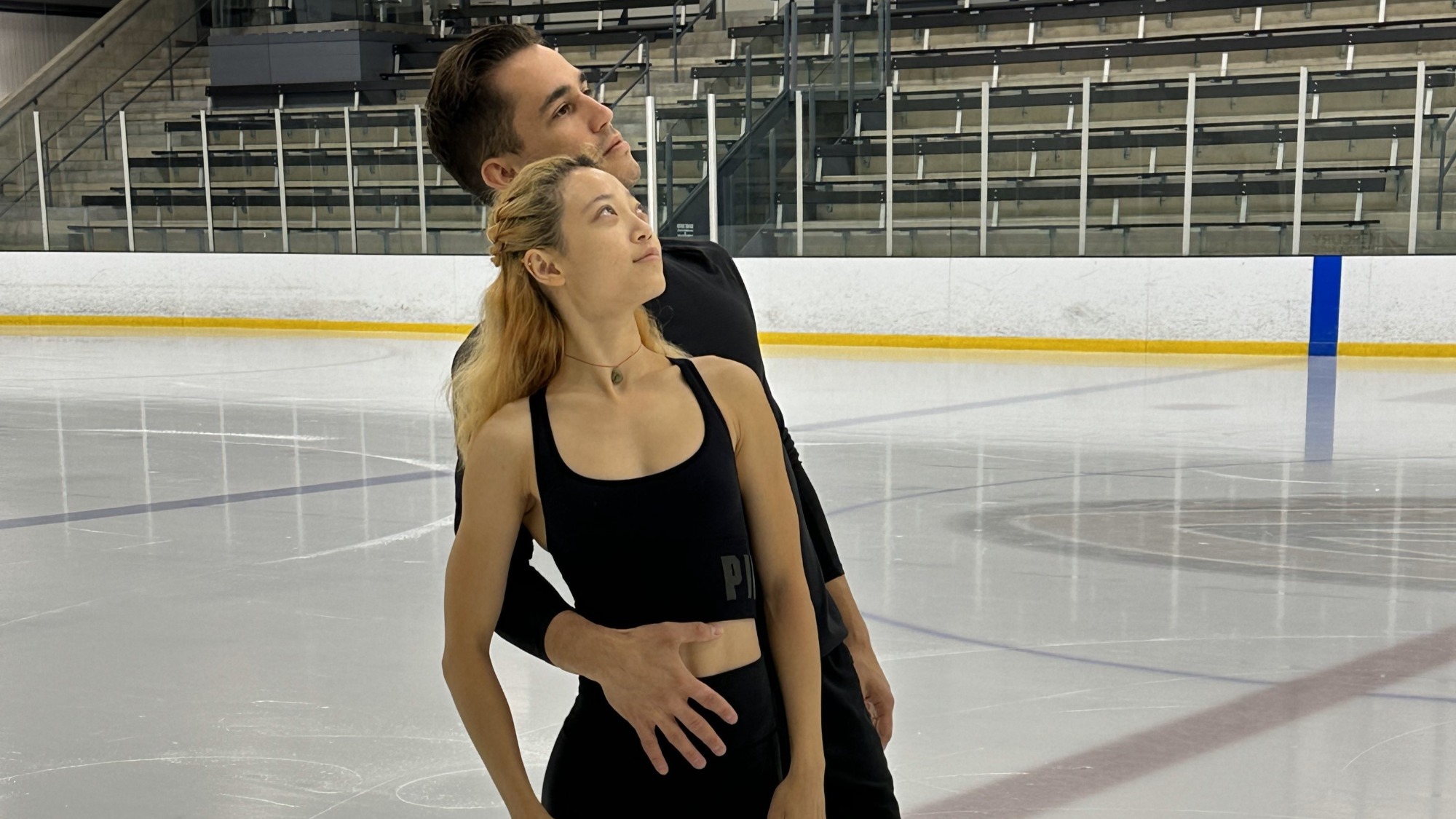 Chelsea Liu stands against Nagy's chest, his hand around her waist. They both look out into the distance, wearing black workout gear. 