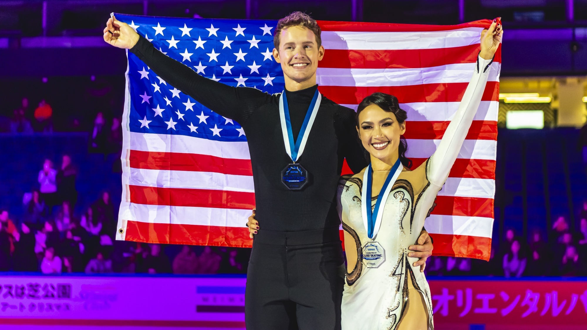 Madison Chock and Evan Bates hold up the USA flag behind them, gold medals around their necks as they smile. He wears all black while she wears a white and beige dress with black trim.
