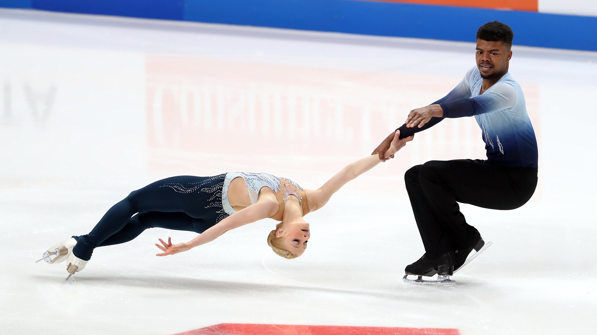 Nica Digerness and Mark Sadusky perform a death spiral. She wears a bodysuit that is naby on the bottom and light blue on top. He wears black pants and his shirt is ombre, from light blue at the top to black at the bottom.