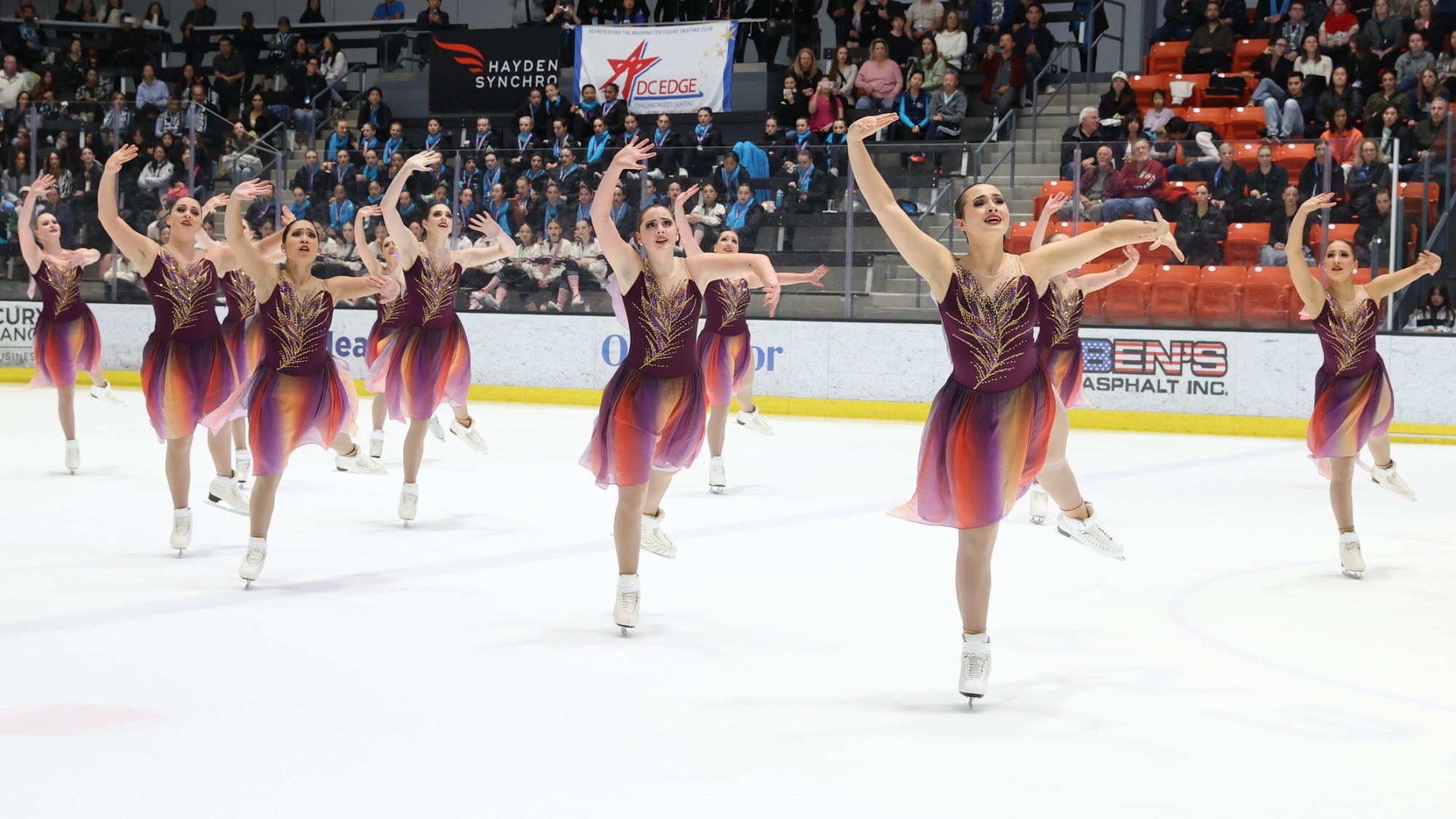 The Haydenettes, in dresses with a purple top and leaf colored pattern in gold that goes up the bodice of the dress and skirts that are multicolored with purple, red and gold, skate toward the camera in unison, their left arm up toward the sky and right arm out to the side.