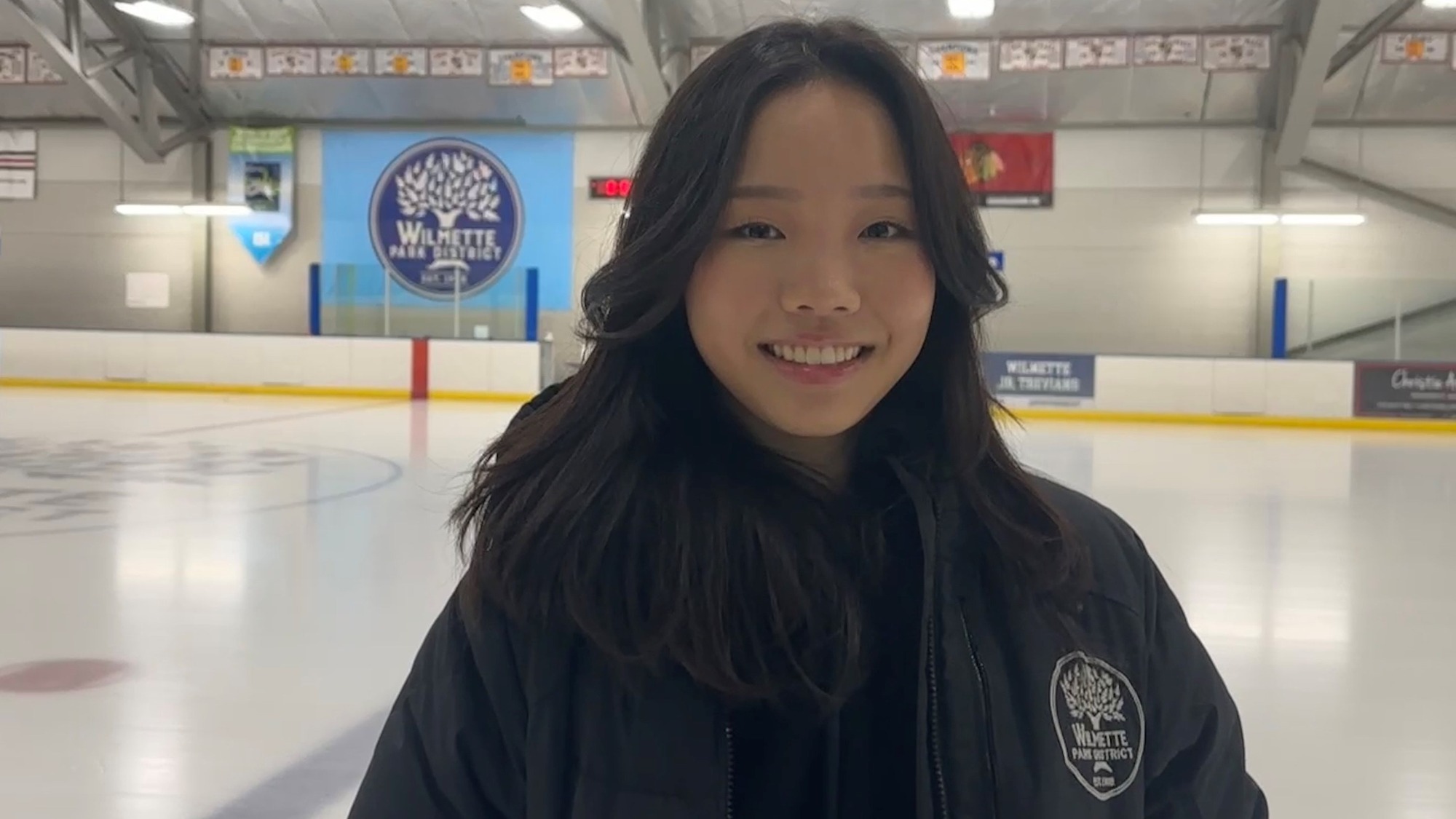 Amy Ji, wearing a black sweatshirt, smiles for the camera. In the background is an ice rink.