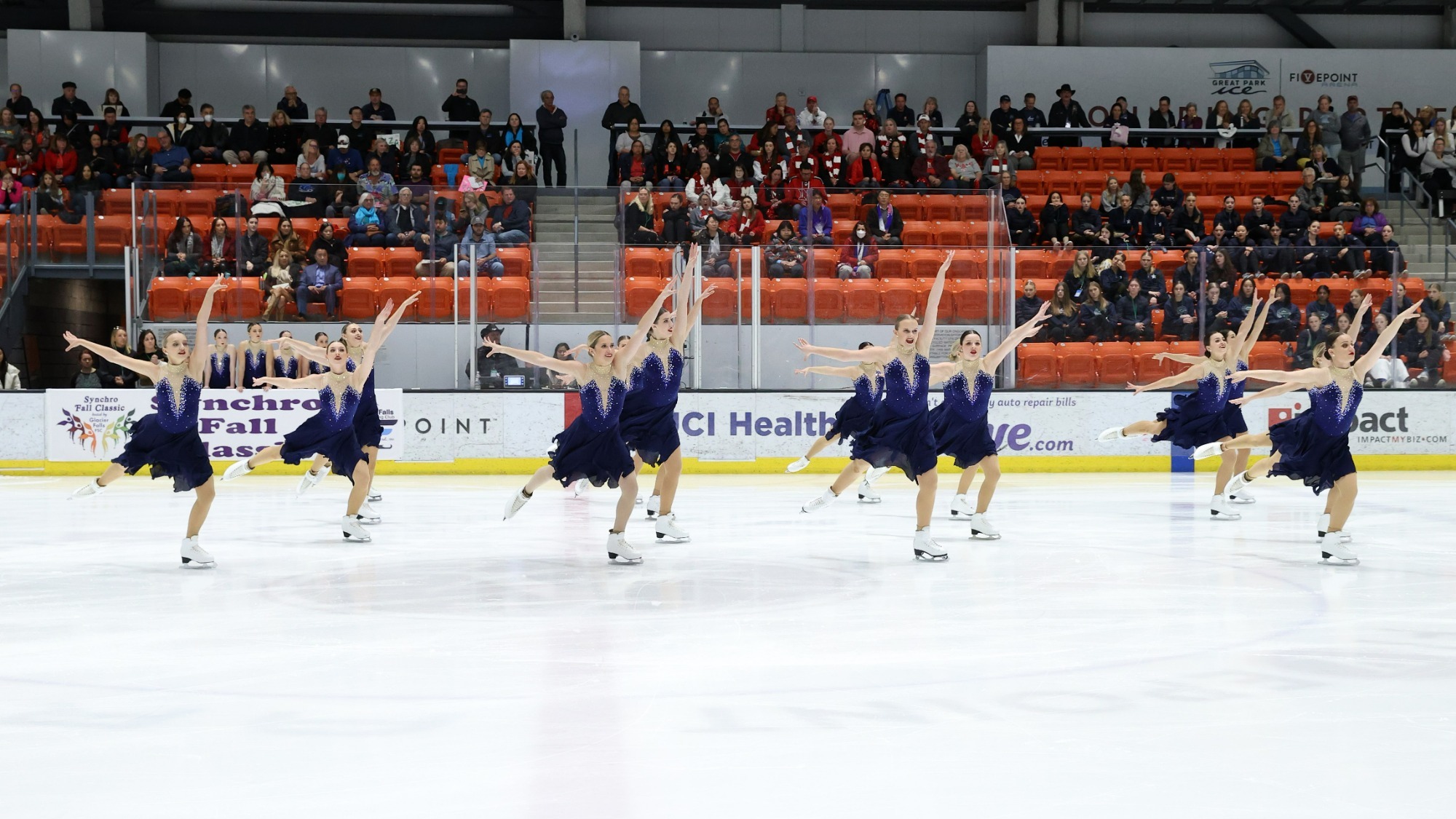 Photo of Adrian College competing on the ice.The group of female skaters stands with their arms outstretched with their right legs extended behind them. They are all wearing dark purple dresses with their hair in tight buns. Fans sit in the stands looking on in the background. 
