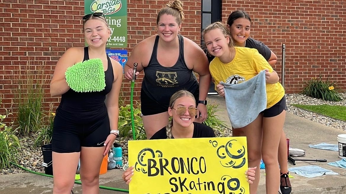 Photo of five female members of the Western Michigan University Figure Skating Team smiling for a photo at a car wash fundraiser. Four are standing and are wearing tank tops and shorts with one kneeling in front holding a sign that says 