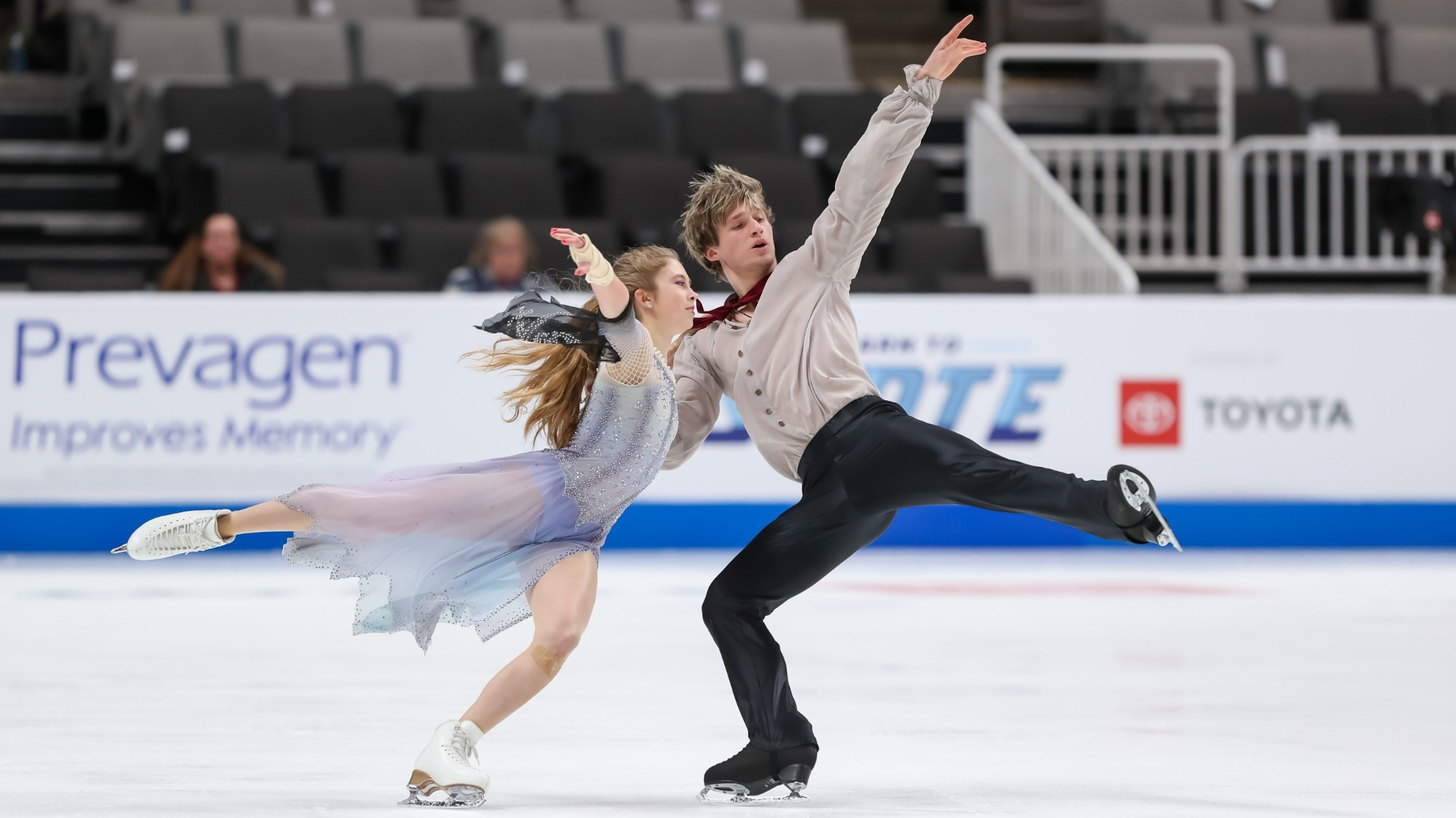 Leah Neset and Artem Markelov compete in the free dance at the 2023 U.S. Figure Skating Championships. Leah (Left) is wearing a light blue skating costume. She has long blonde hair she is wearing down. Artem (right) has short blonde hair and he is wearing a cream colored button up shirt with black pants. Both are skating with one arm and leg extended outward. 