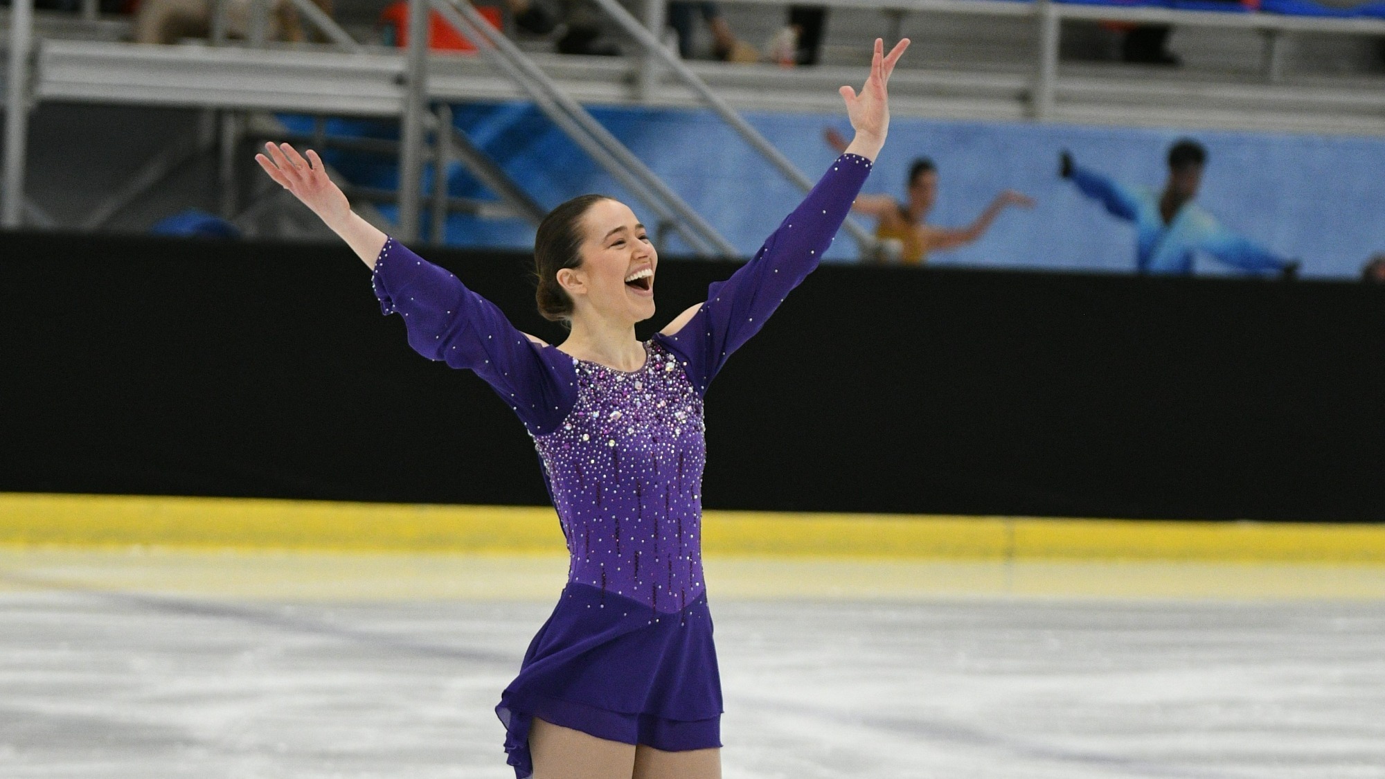 Anya Saretzky has a big smile on her face as she acknowledges the crowd at the 2022 U.S. Adult Figure Skating Championships. Anya is a white woman with brown hair tied back in a bun. She is wearing a purple long sleeve and sparkly skating costume. Her arms are lifted above her head