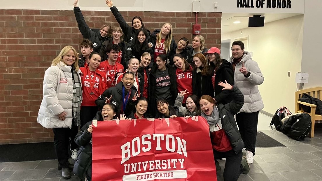 The Boston University figure skating team poses against a brick wall with the Boston University figure skating flag, taking a silly picture with fun looks on their faces.