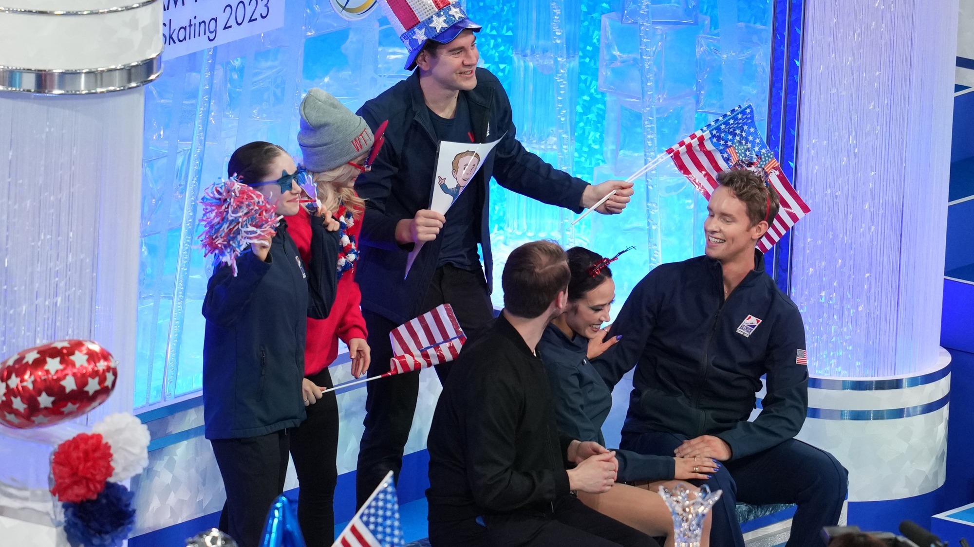 Madison Chock and Evan Bates smile next to their coach as they receive their rhythm dance scores. Alexa Knierim and Brandon Frazier and Isabeau Levito stand behind them, decked out in USA themed gear.