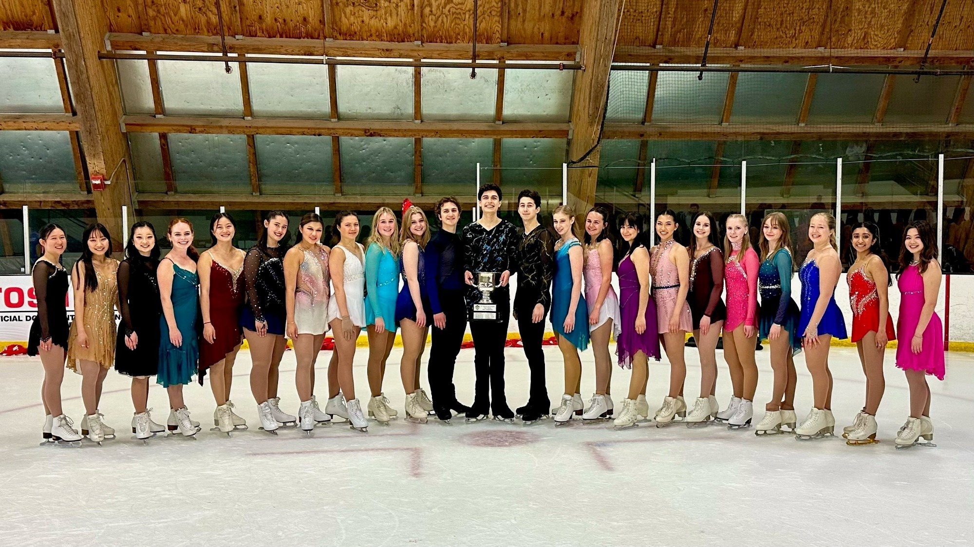 The Boston University Figure Skating Team poses with their first place trophy at the National Intercollegiate Final in Anaheim, California. All of the skaters are wearing various colored free skate costumes 