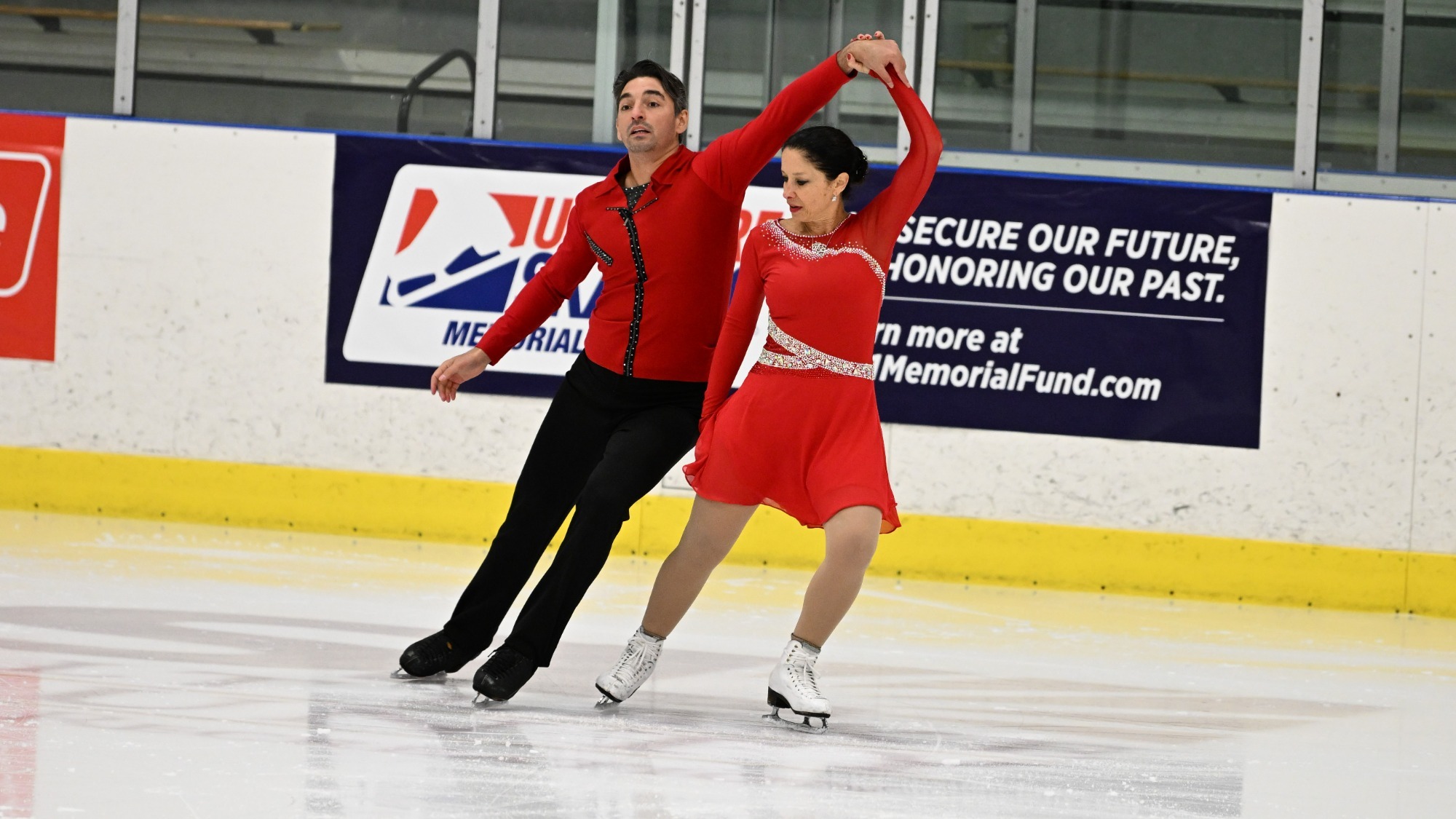 A male and female skater compete at the 2023 U.S. Adult Figure Skating Championships. The woman is wearing a red long sleeve skating costume and the man is wearing a red long sleeve shirt with black pants.