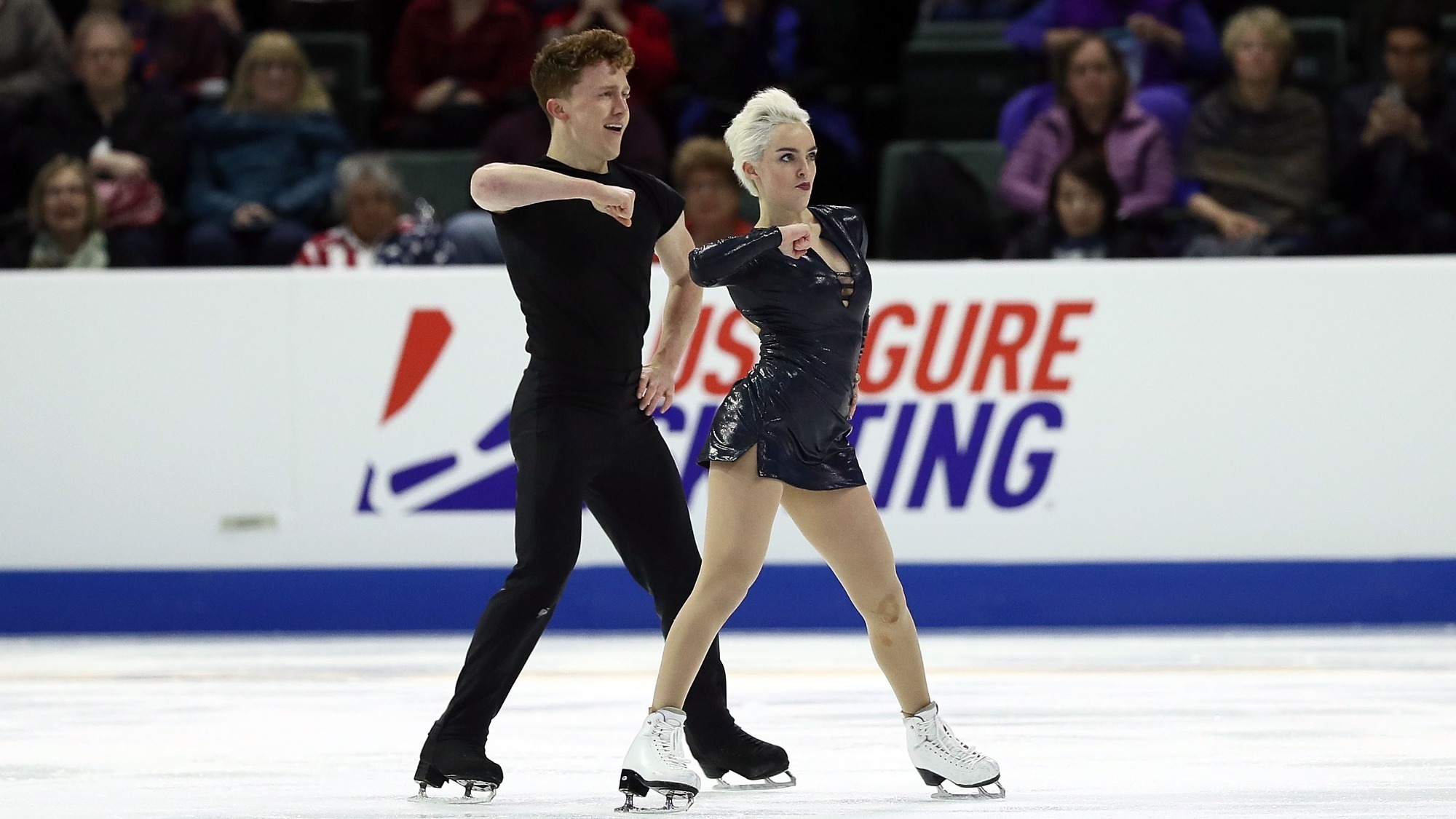 Karina Manta stands in front of her partner, Joe Johnson, during a performance at Skate America. They both wear black and are performing a dance move in synch, their left arm horizontal at chest level.