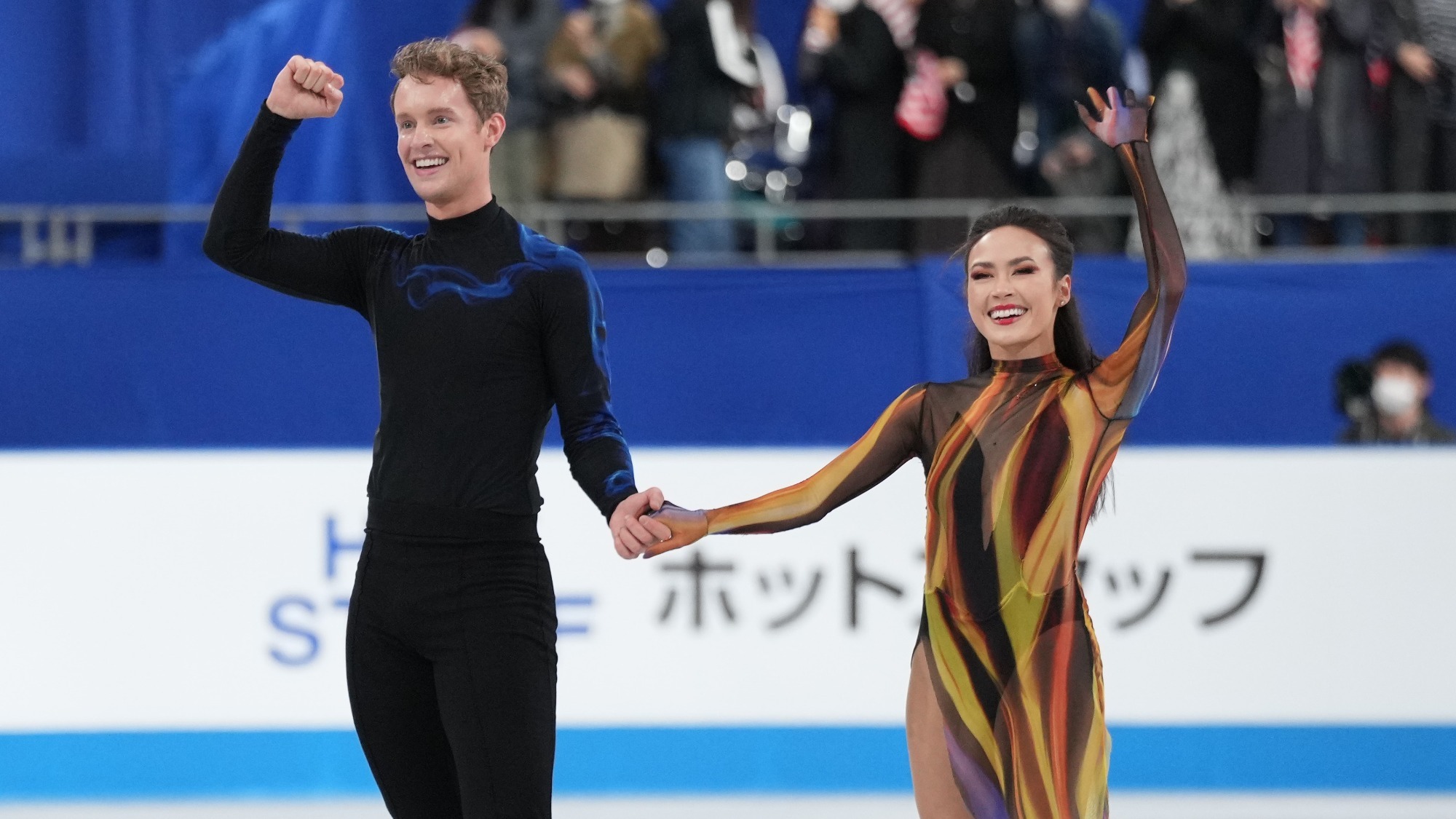 Madison Chock and Evan Bates, during their free dance bow at World Team Trophy. They hold hands. She waves with her other hand while he pumps his fist. 