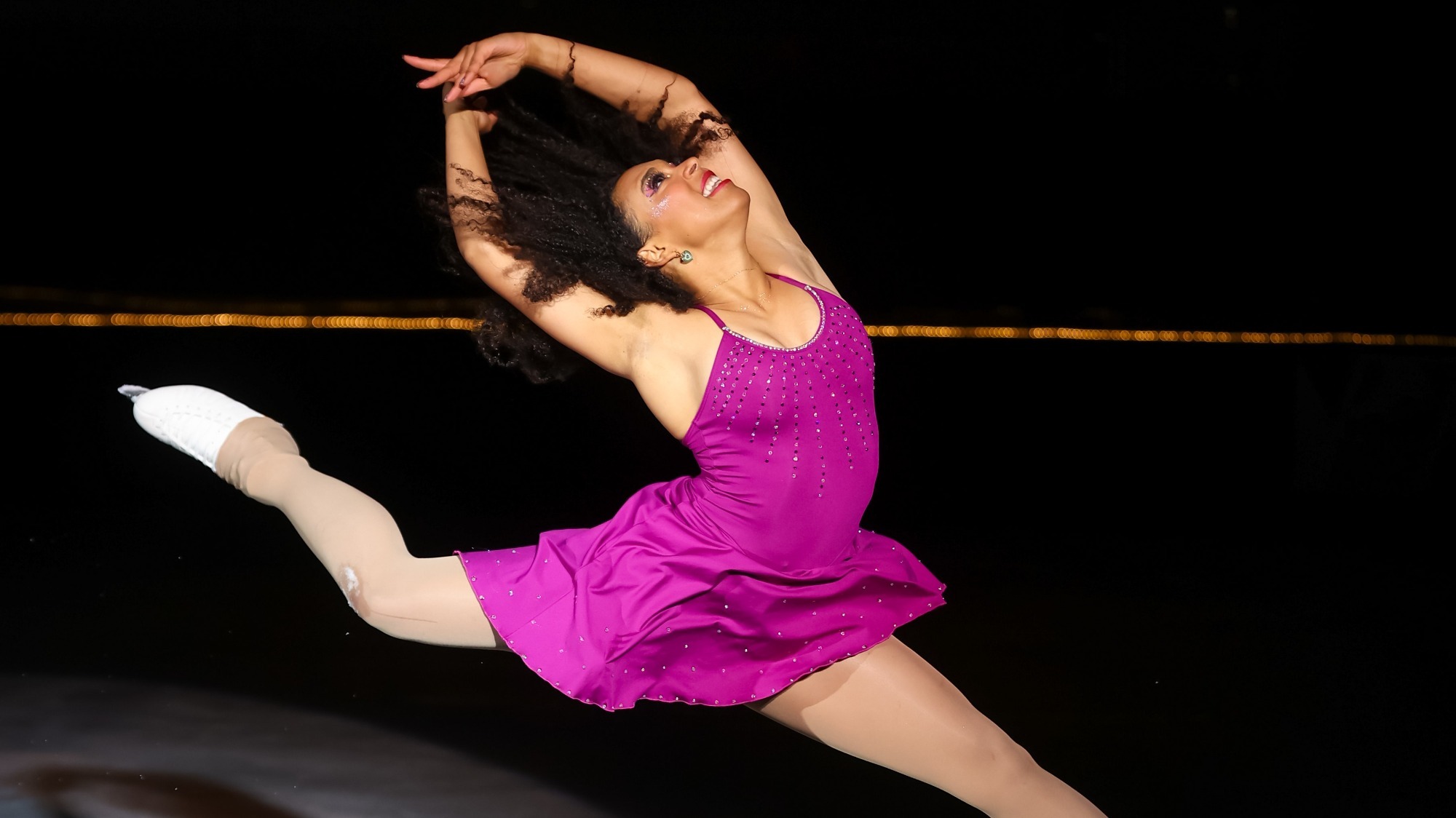 A female skater with long black hair and a purple skating costume leaps in the air with her arms above her head