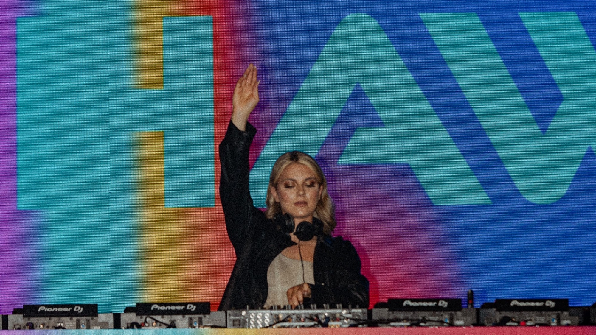 Kaitlin Hawayek DJs at the Grand Prix party, mixing with one hand and one hand in the air. She wears a black leather jacket, and the first three letters of her latst name - HAW appear on a rainbow screen behind her.