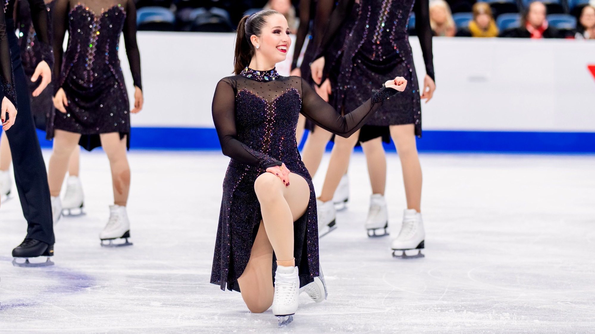 Elissa Ekempisty hits a pose as a member of the Haydenttes at the World Championships during the short program, her knes and back leg on the ice as she kneels, a big smile on her face. She wears a sparkly black dress.