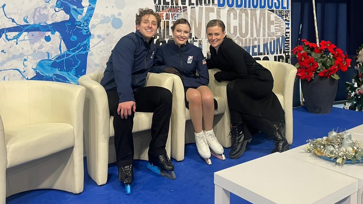 Madison Hubbell (right) sits in the Kiss and Cry with Christina Carreira and Anthony Ponomarenko. Carreira and Ponomarenko are wearing their USA jackets over their free skate costumes and their skates while Hubbell wears a long black coat with black pants and boots..