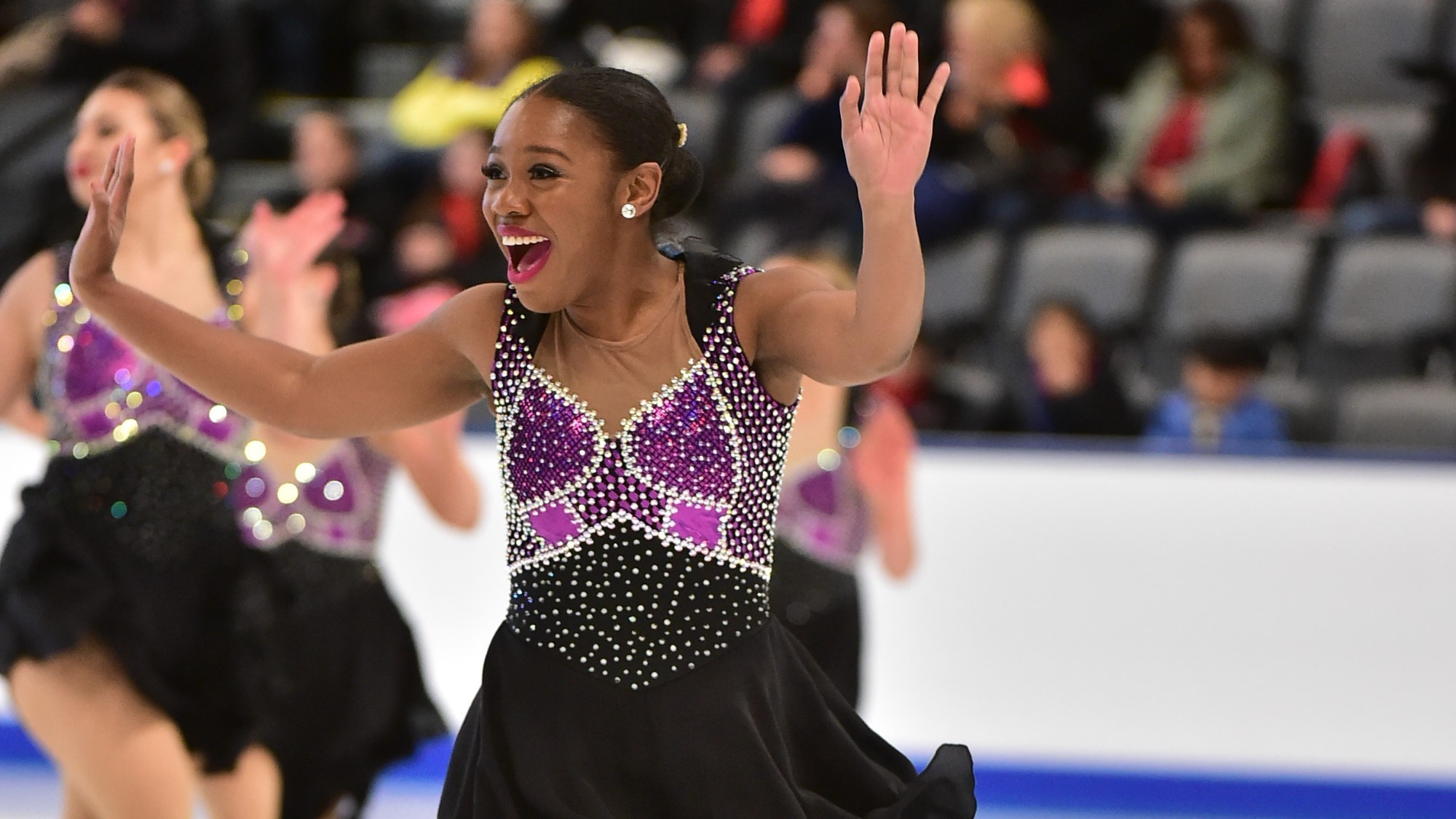 Leatrice Bulls, in a black dress with sequins from the waist up and purple on the chest, holds her hands up with a very expressive, excited look on her face.