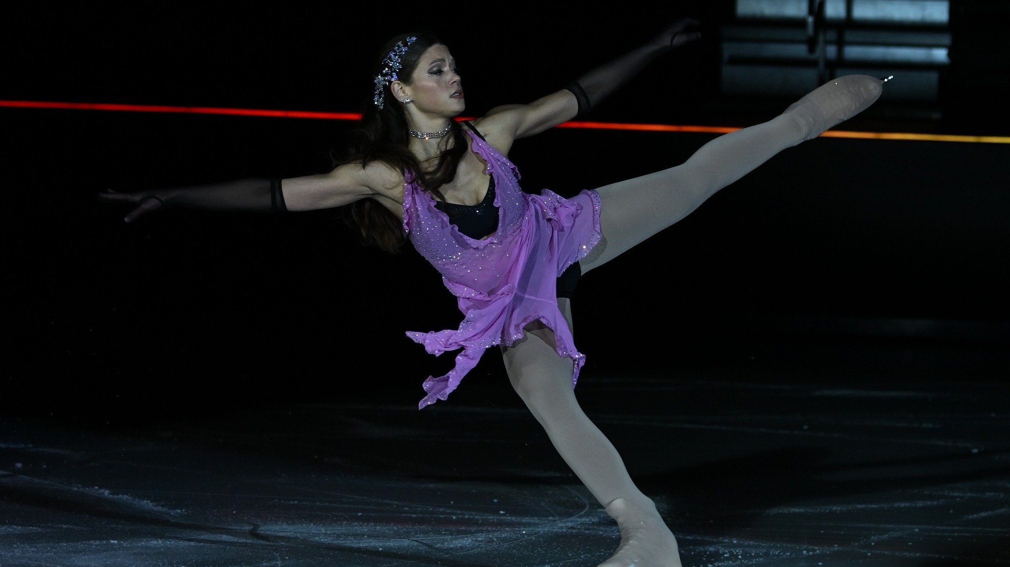 Ashlyn Olson competes in the senior Parade of Champions at 2023 National Showcase. She is a young woman with long black hair. She is wearing a purple skating costume with black gloves and a jeweled headpiece. She stands with both arms and one leg extended outward