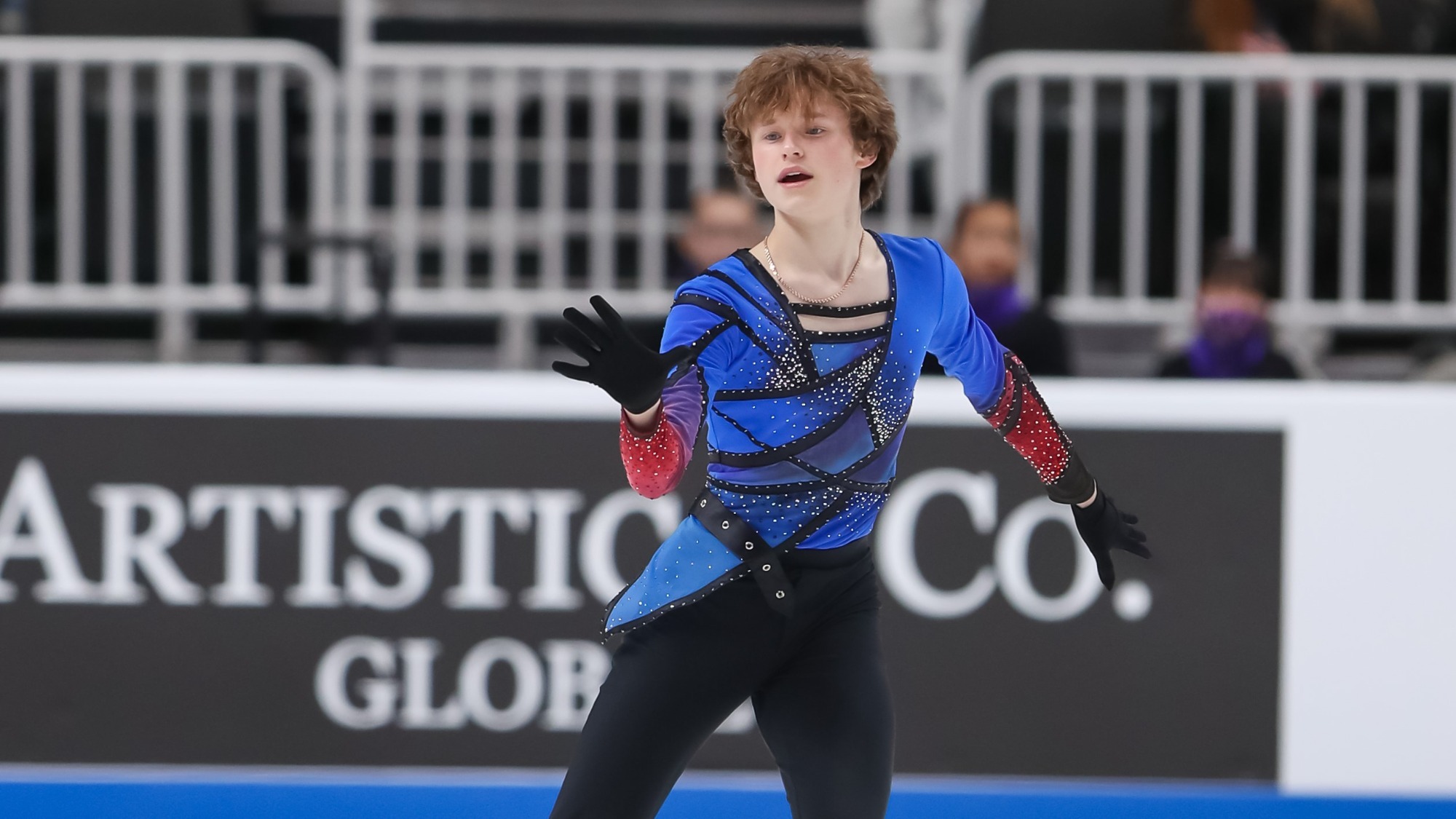 Ilia competes in the men's free skate at the 2022 U.S. Championships. Ilia is a young man with short brown hair wearing a long sleeve red and blue top with black pants. He holds one hand out in front of his body. 