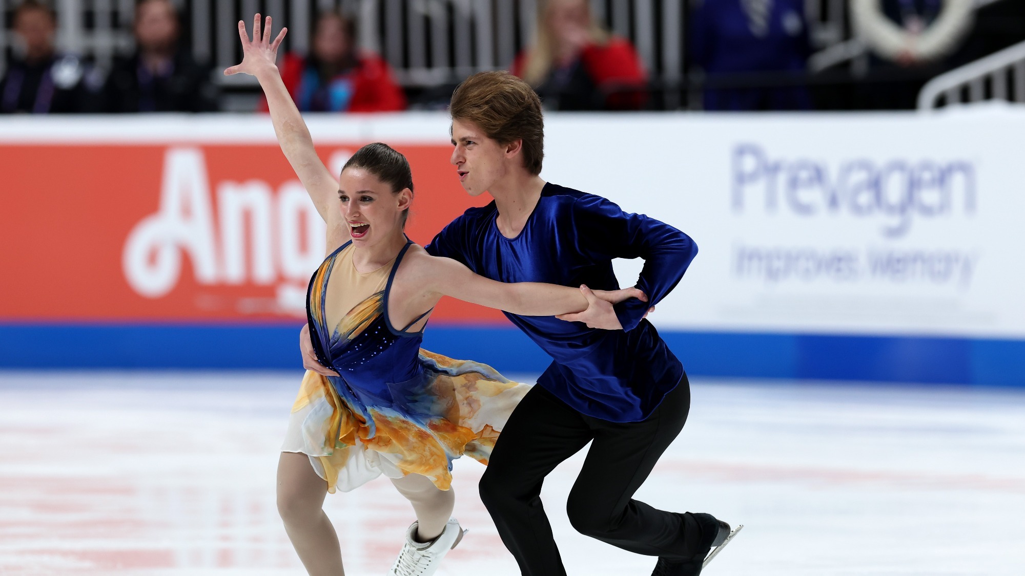 Gage Brown holds Oona' Brown's hand and her waist, as they skate. He has her free hand up in the air and an excited look on her face, wearing a blue, orange and white dress. He wears black pants with a navy blue top.