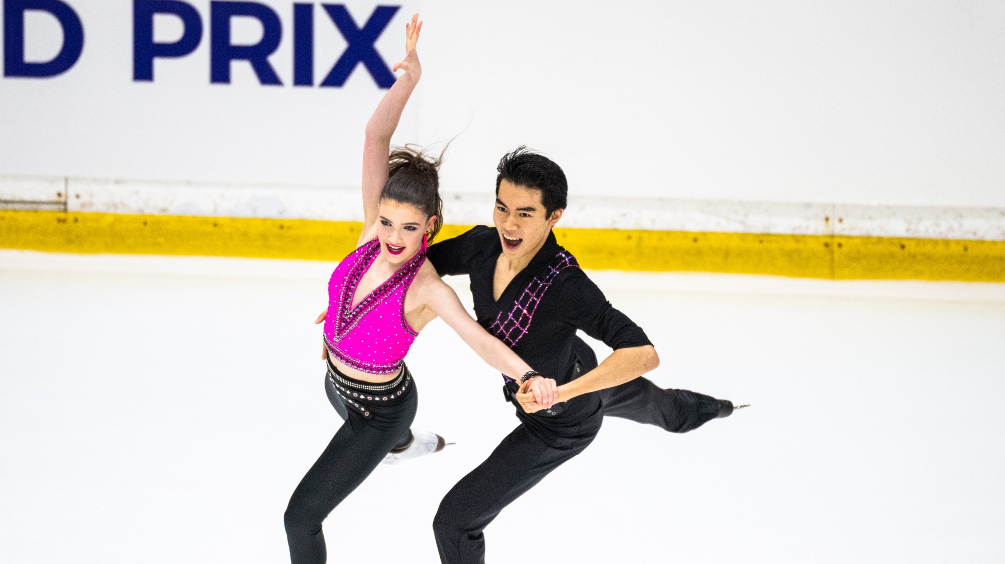 Yahli Pedersen and Jeffrey Chen perform to their 80s style rhythm dance. Both with joyful expressions, Pedersen wears a bright pink top and leather pants while Chen wears black pants and a black top with a pink rhinestone detail.