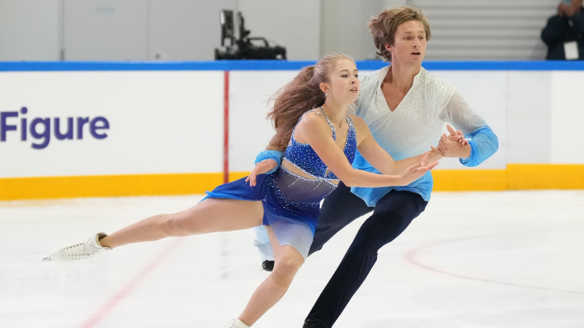 Leah Neset and Artem Markelov skate at the 202 Junior Grand Prix Thailand. They are skating side by side holding each other's hand. Both have one leg extended behind them. 