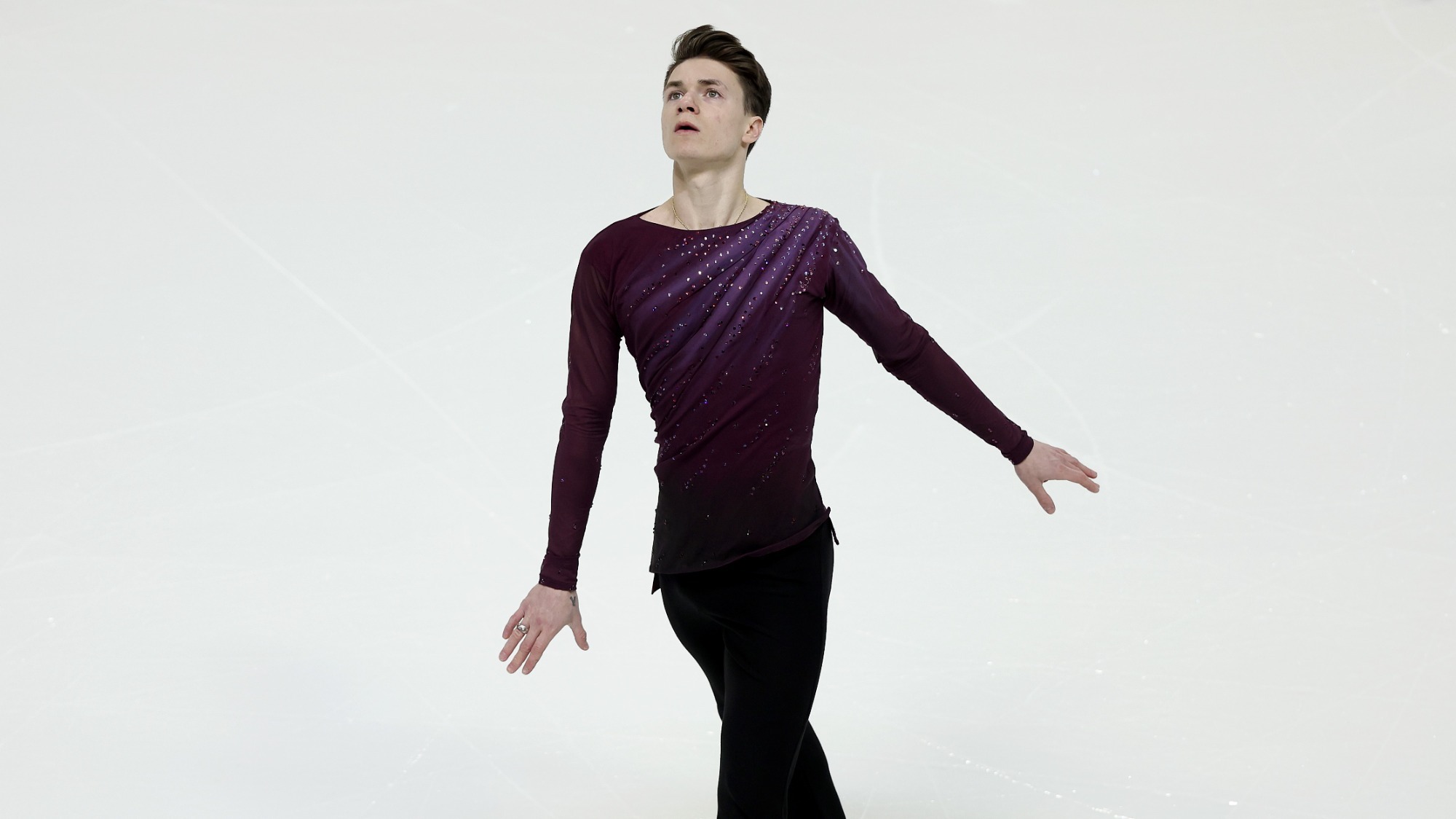 Maxim Naumov, in a purple top that has jewels running diagonally and black pants, stairs up toward the sky with a pensive look on his face.