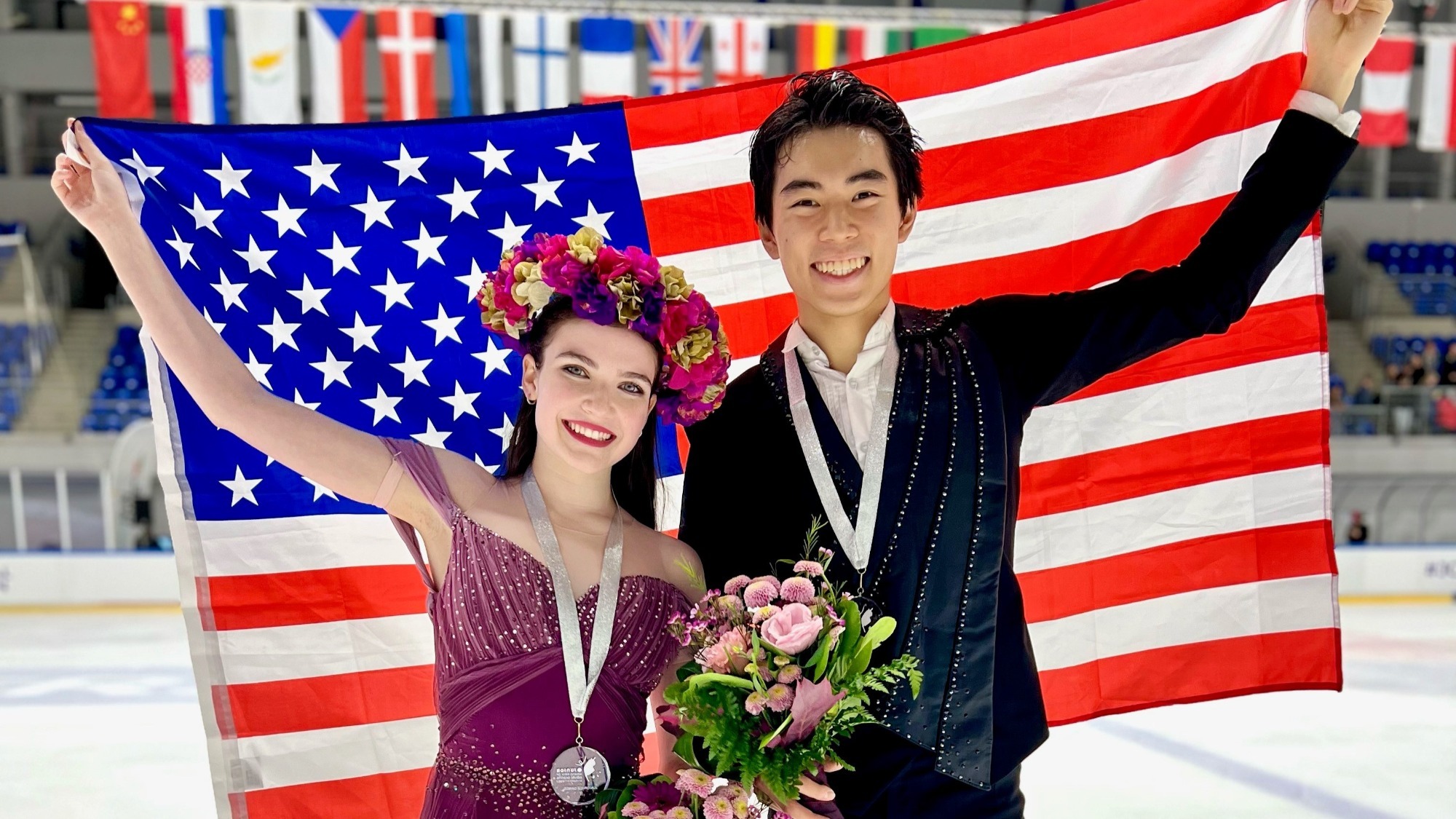 Yahli Pedersen and Jeffrey Chen hold up the U.S. flag behind them, silver medals around their necks/ She wears her purple free dance dress, a flower crown around her head, while Jeffrey wears a black jacket with jewels down the collar.
