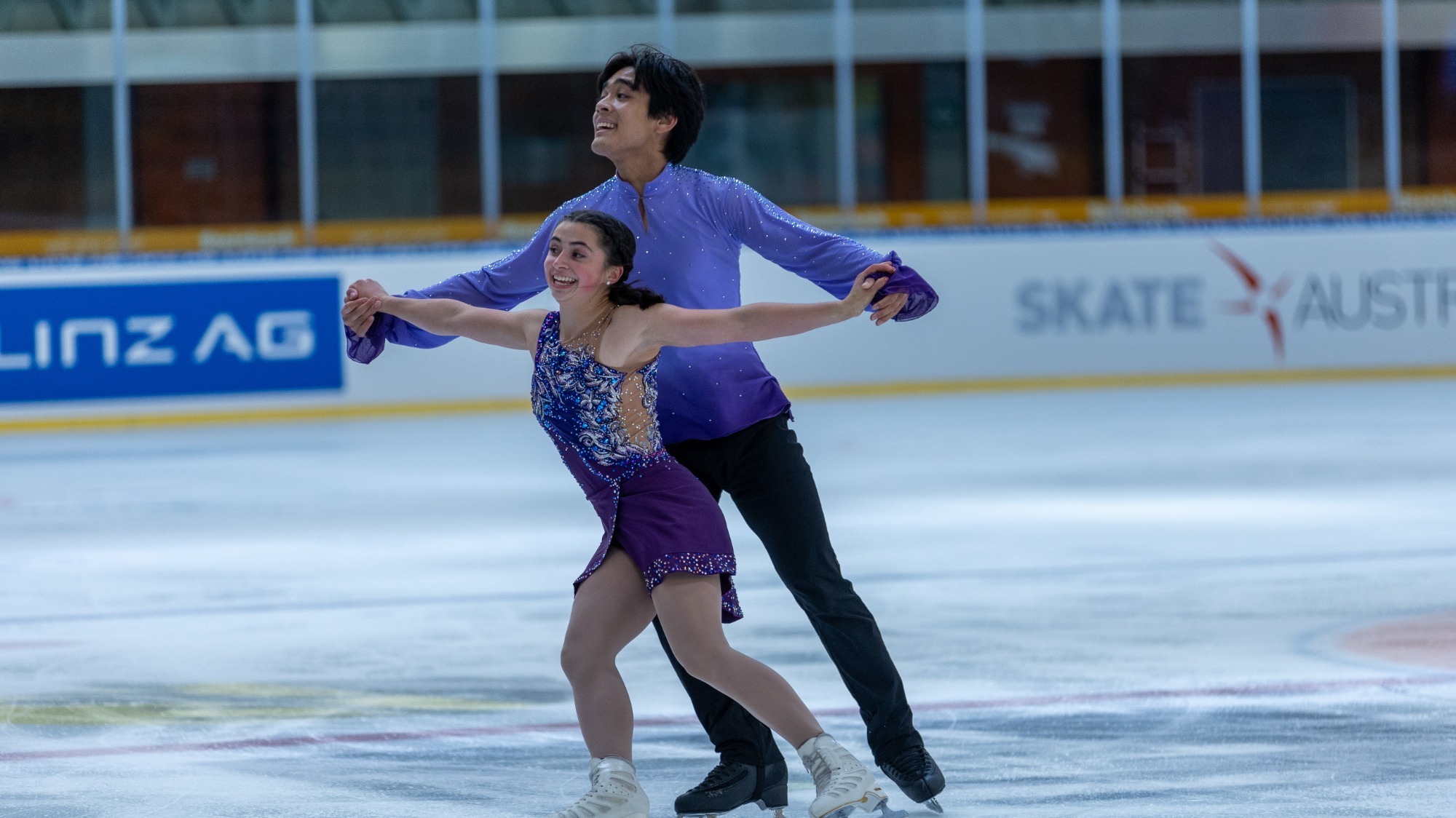 Olivia Flores and Luke Wang compete at JGP Austria. Olivia stands in front of Luke with her arms outstretched at her sides. She is a short woman with short black hair tied back into two braids and is wearing a dark blue and jeweled skating costume. Luke stands behind Olivia holding both of her hands. He is much taller with short black hair and is wearing a blue long sleeve top and black pants 